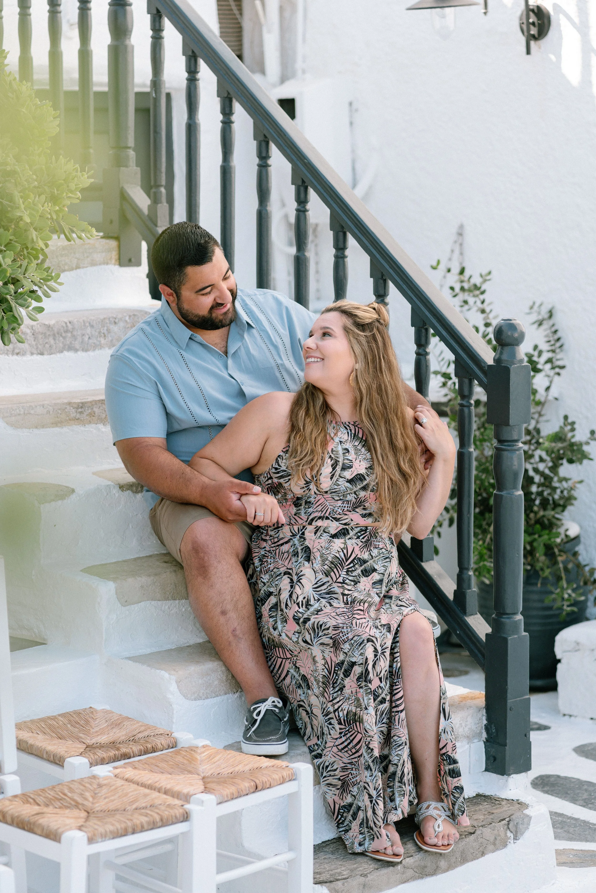 A couple sits on white stairs outside, smiling at each other. The man is wearing a light blue shirt and khaki shorts, the woman a floral dress. They are holding hands, and the scene has greenery and a black railing.