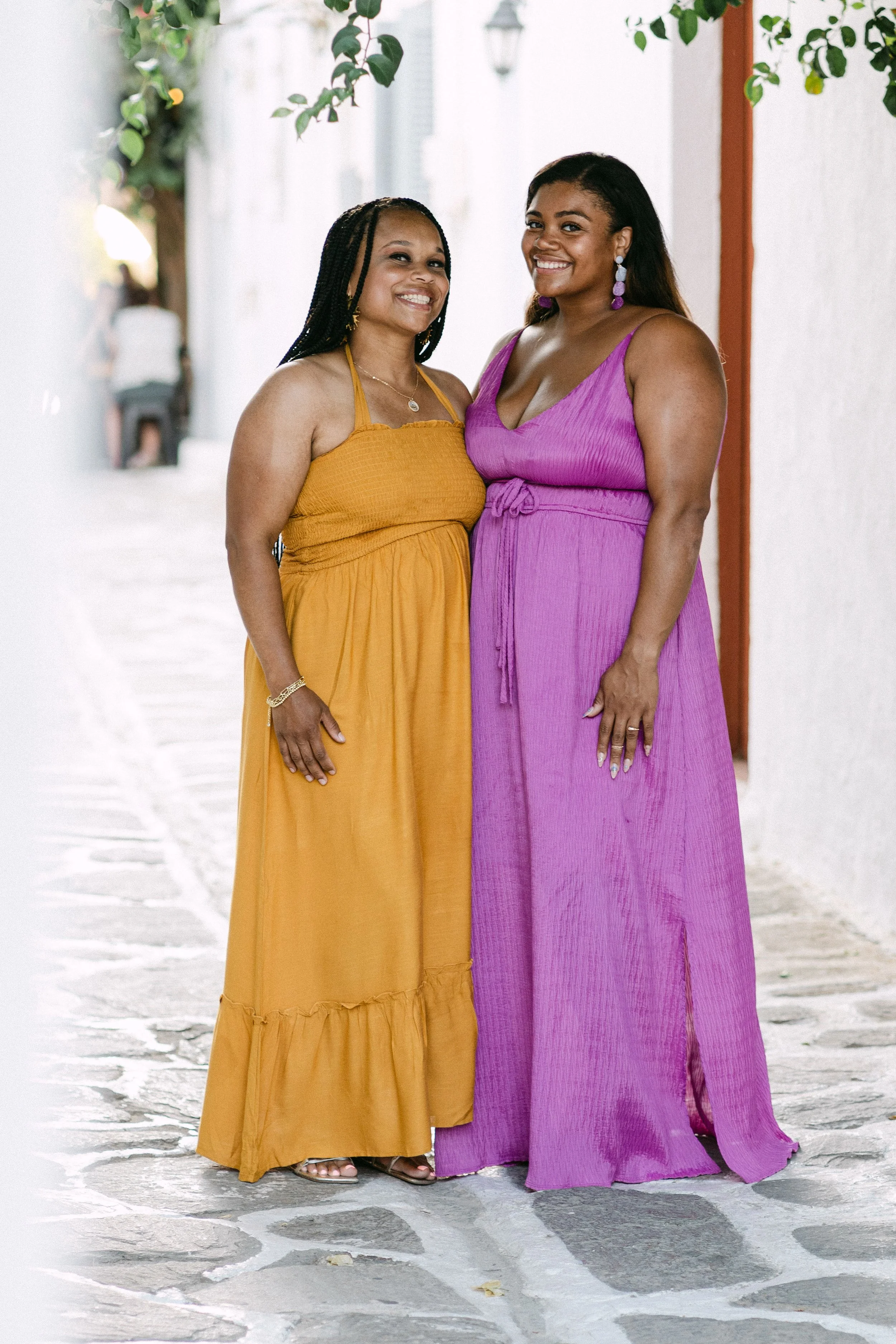 Two women standing together outdoors, smiling, with white walls and leafy branches overhead. One woman wears a yellow dress, the other a purple dress, both with jewelry and relaxed poses.