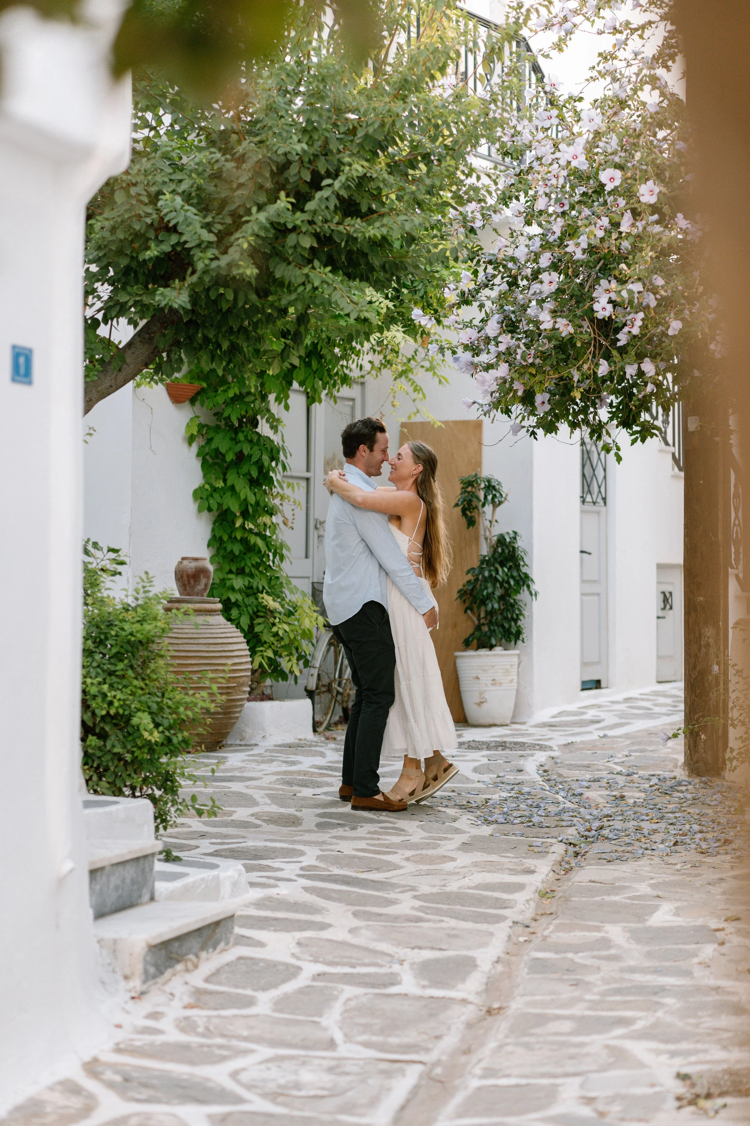 A couple dancing together outdoors on a cobblestone path surrounded by white buildings and lush greenery, with a flowering tree overhead.
