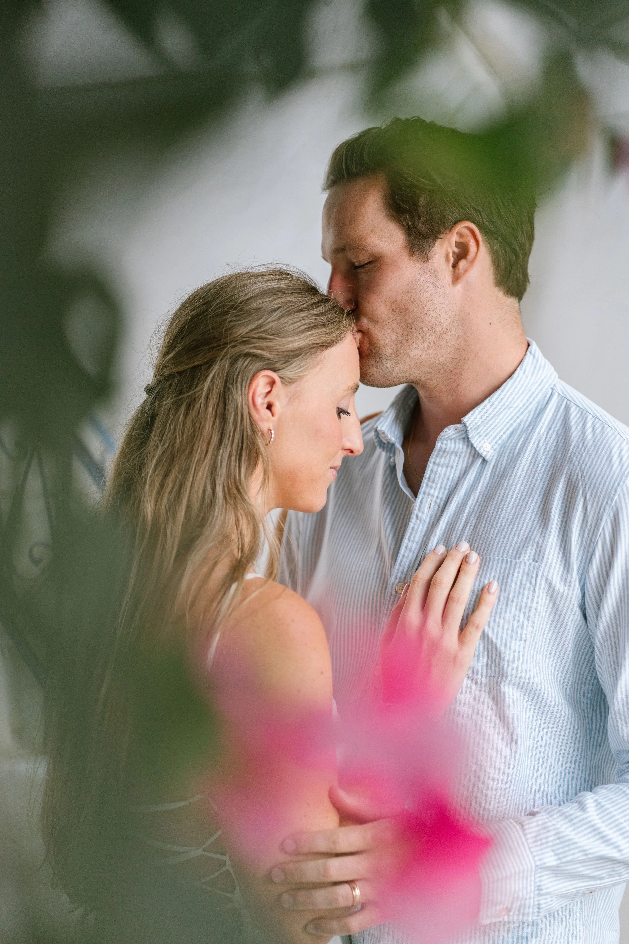 A man kisses a woman on the forehead while she closes her eyes, both with light skin, partially obscured by blurred green and pink leaves in the foreground.