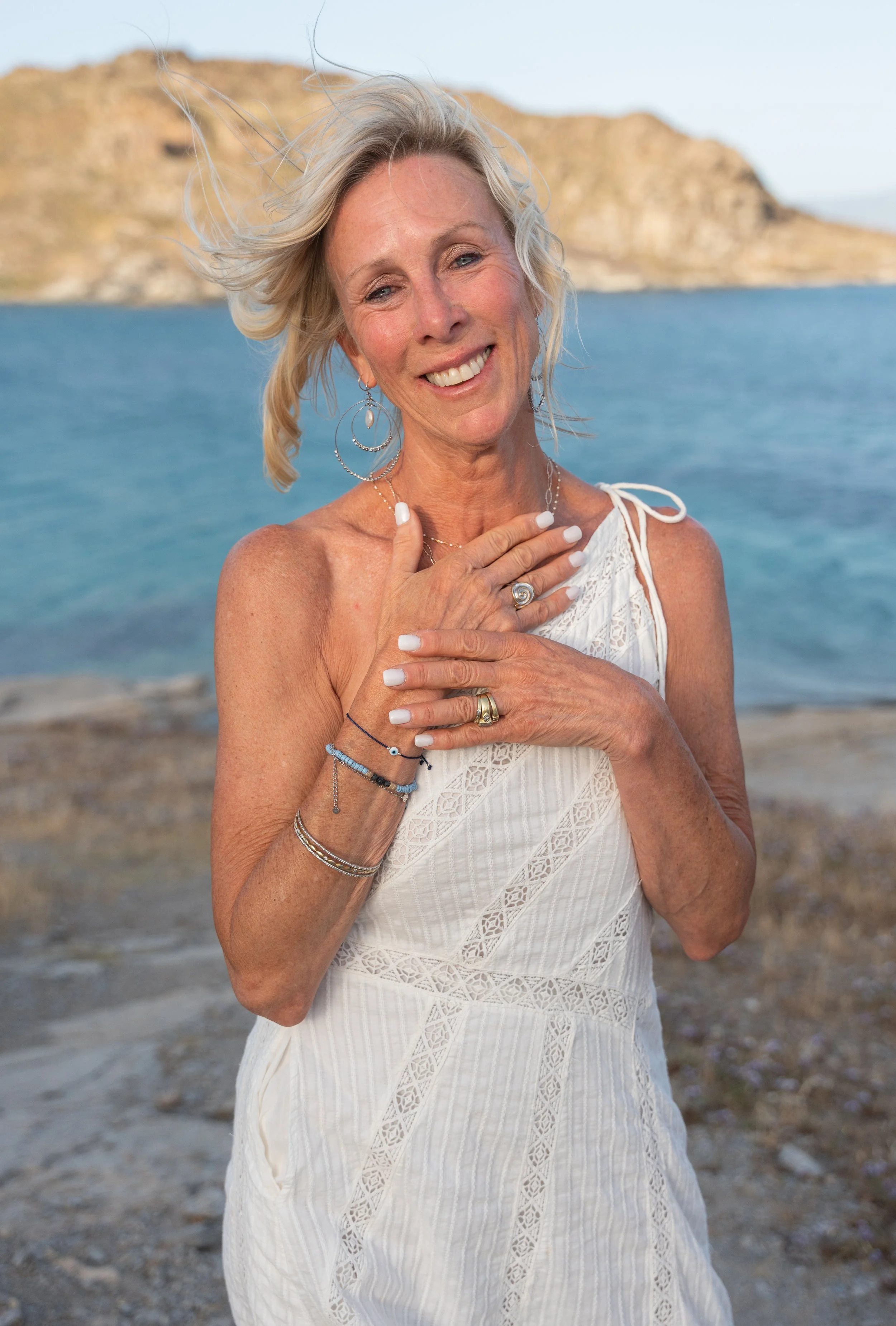 A smiling woman with silver hair, wearing a white dress, standing on a rocky beach near water with cliffs in the background.