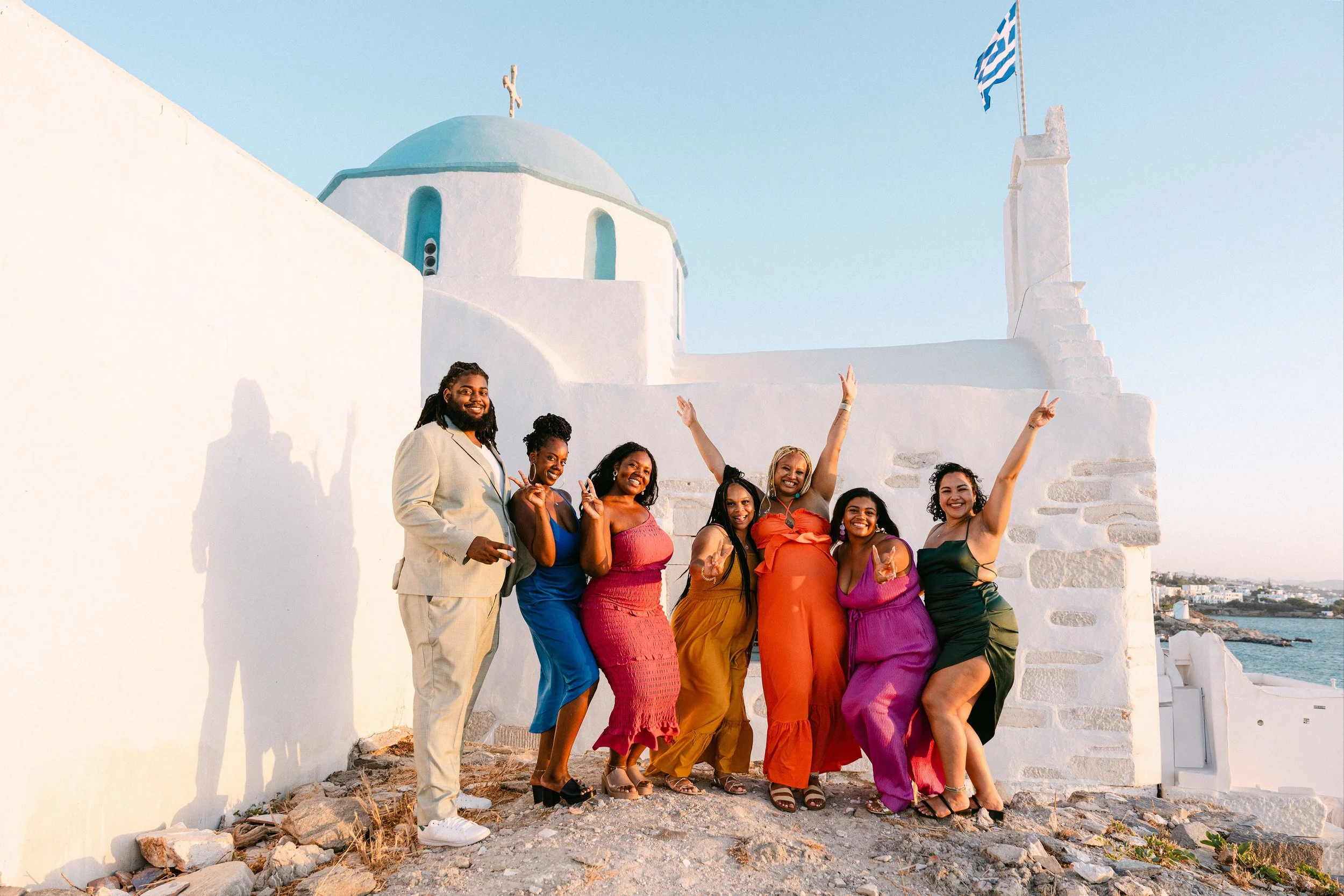 A group of seven diverse people celebrating and posing cheerfully in front of a white Greek-style church with blue domes and flags, near the ocean, during sunset.