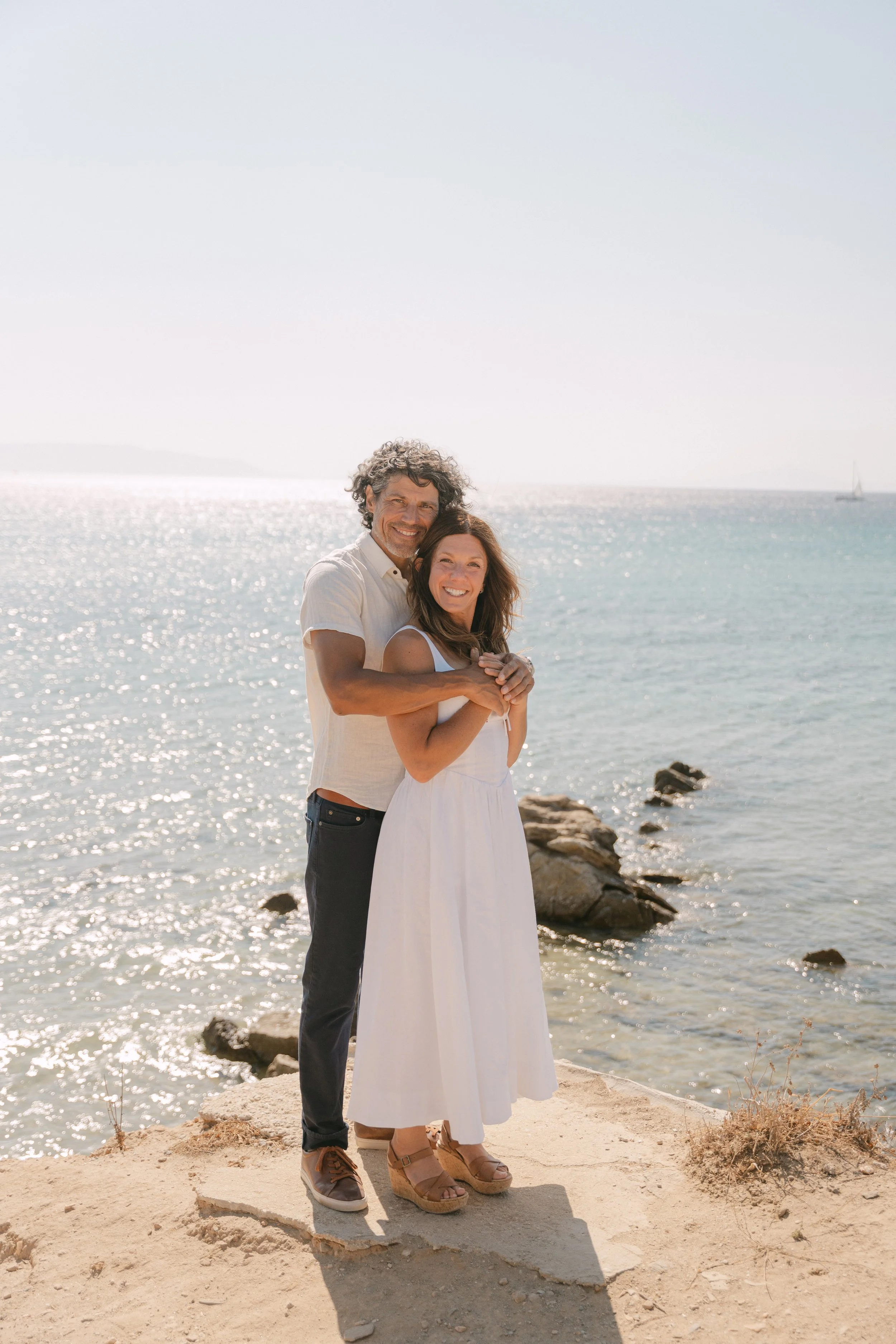 A happy couple standing on a sandy beach, embracing each other, with the ocean and a sailboat in the background, during a sunny day.