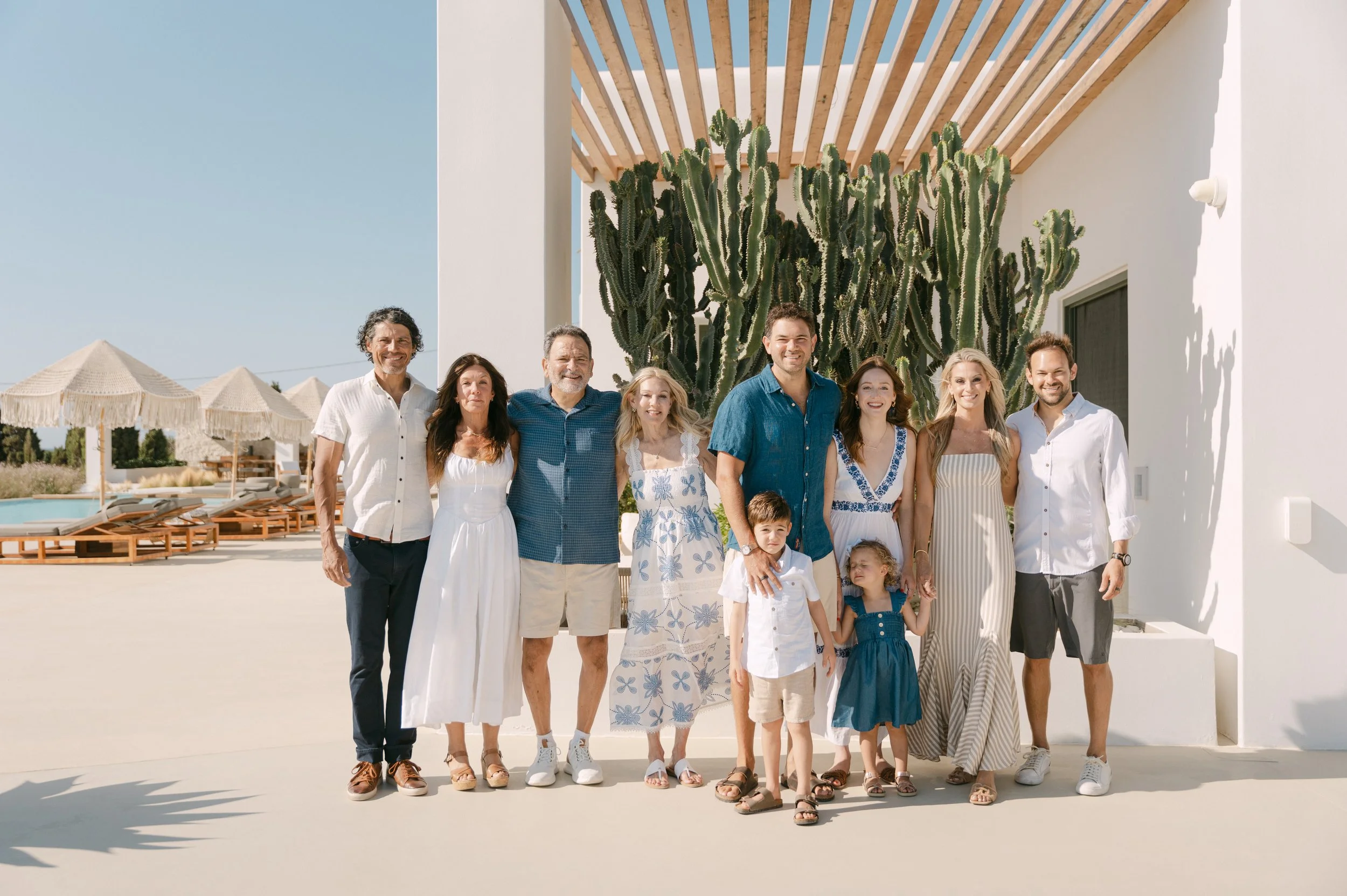 A group of people, including adults and children, standing together outdoors in front of a large cactus plant and a white building, during a sunny day.