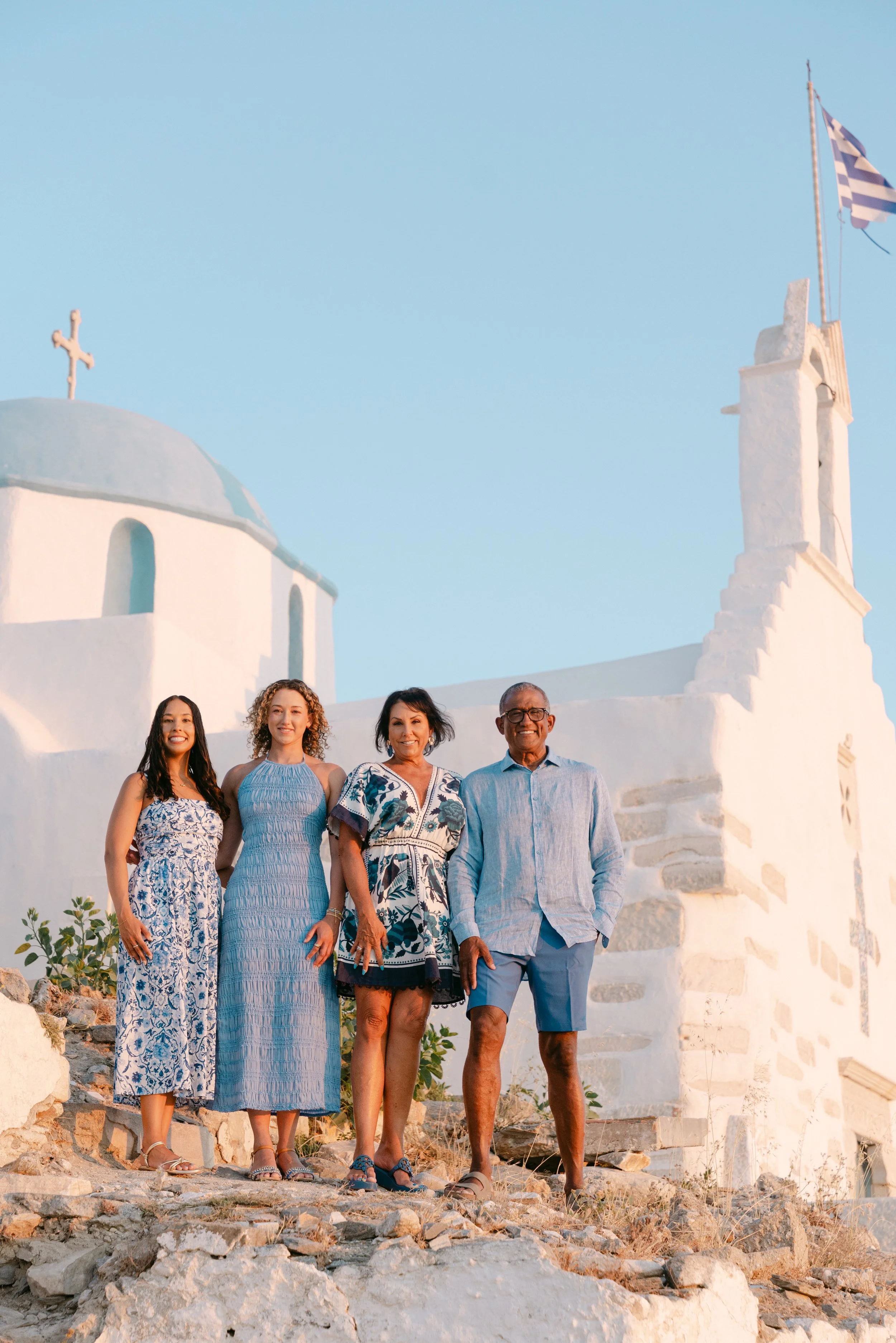 Four people standing outdoors in front of white buildings in a Greek island setting, dressed in summer clothing, with a clear blue sky overhead.