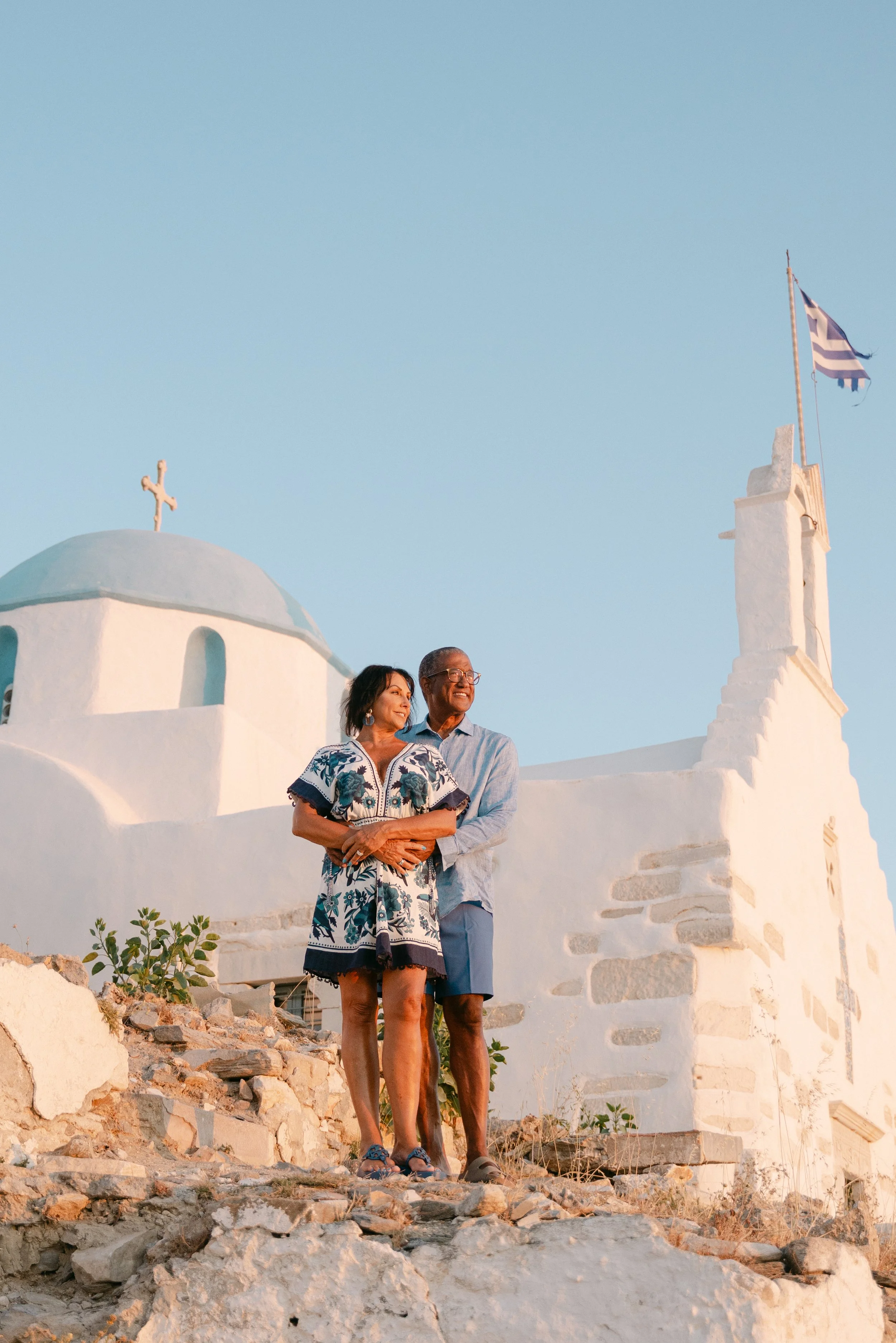 An elderly couple standing on rocky terrain in front of a white Greek church with a dome and cross, and a flagpole with the Greek flag, during sunset.