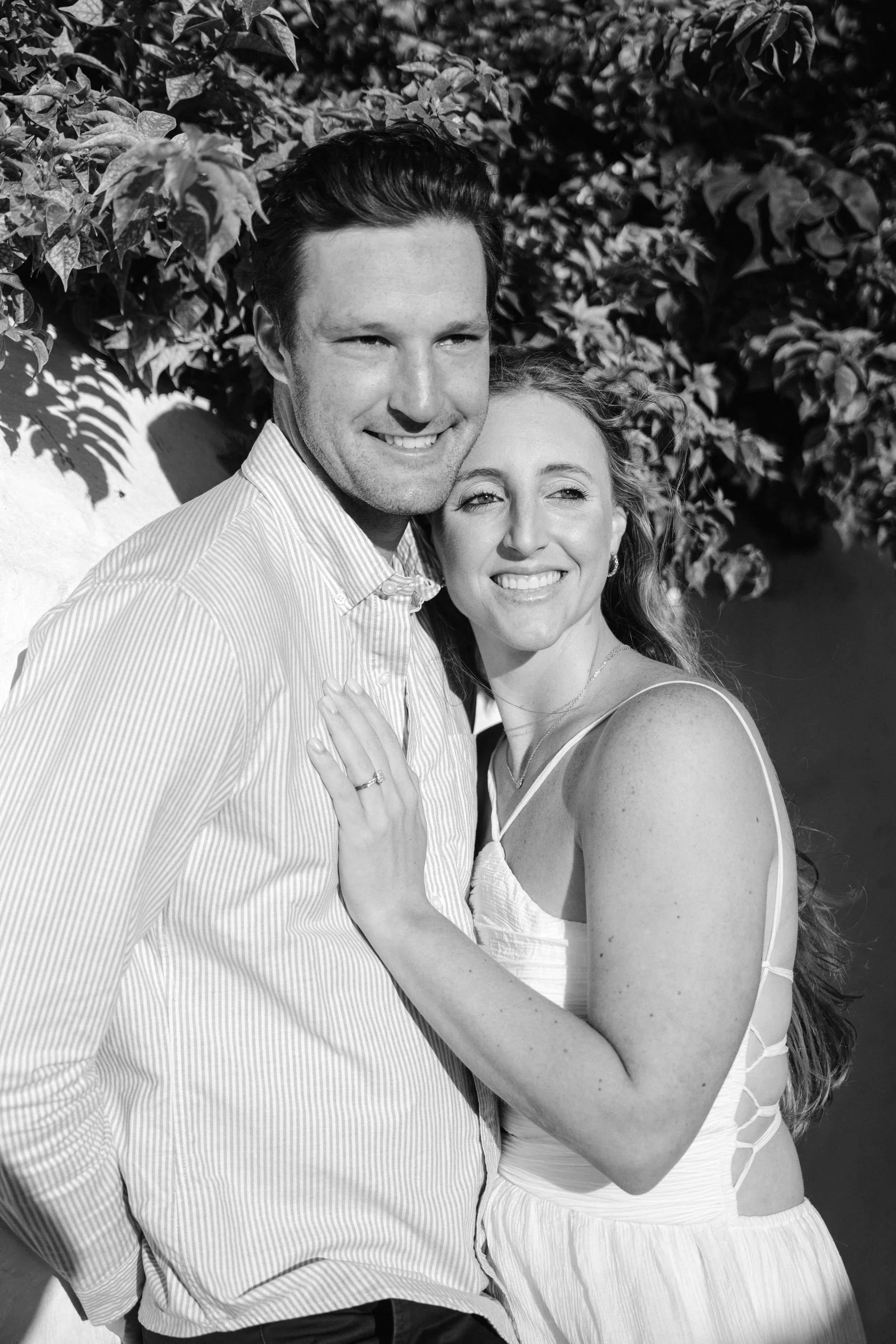 A smiling man and woman embrace outdoors, with leaves in the background, in a black-and-white photo.