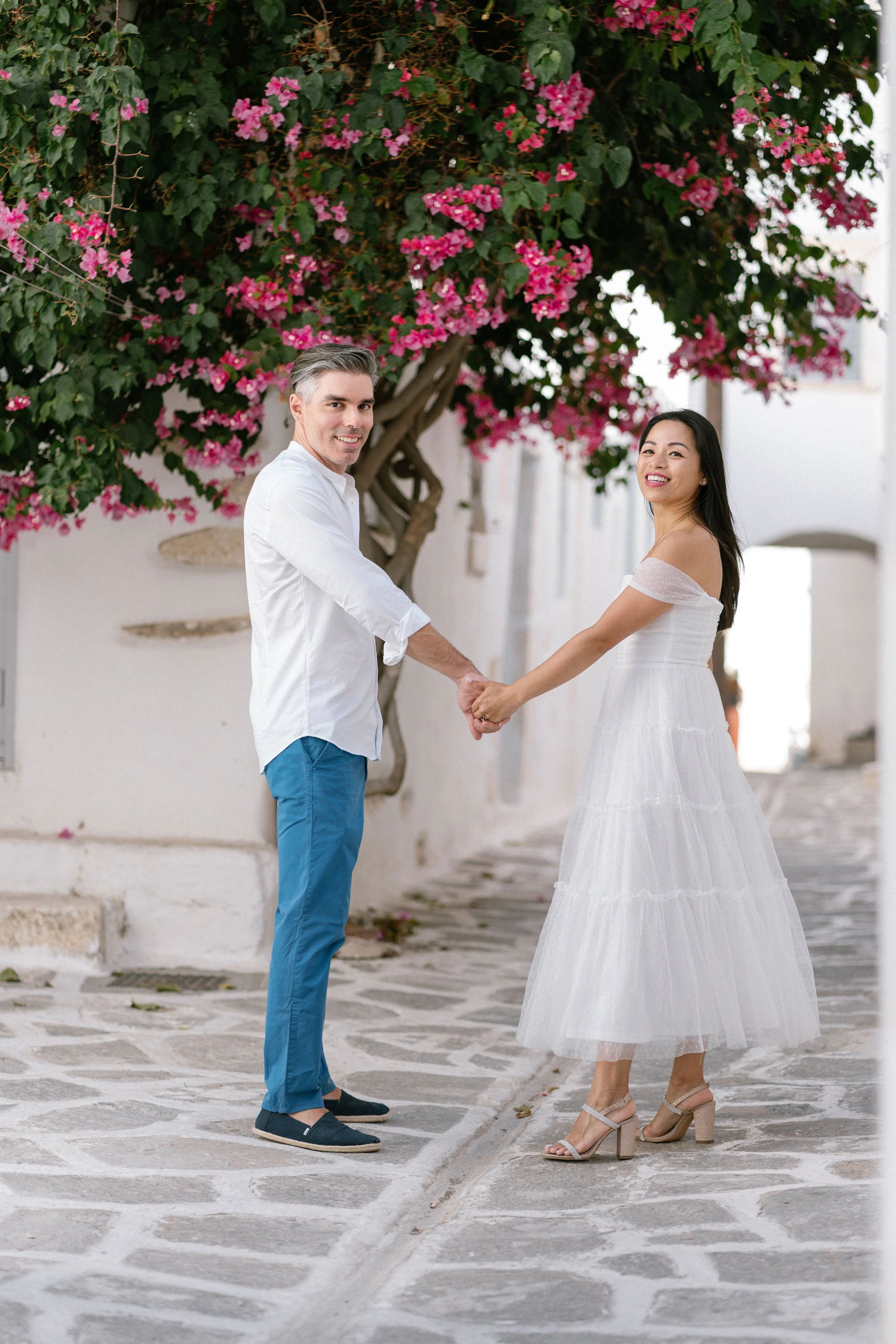 A couple holding hands and smiling on a cobblestone street, with pink bougainvillea flowers and white walls in the background.