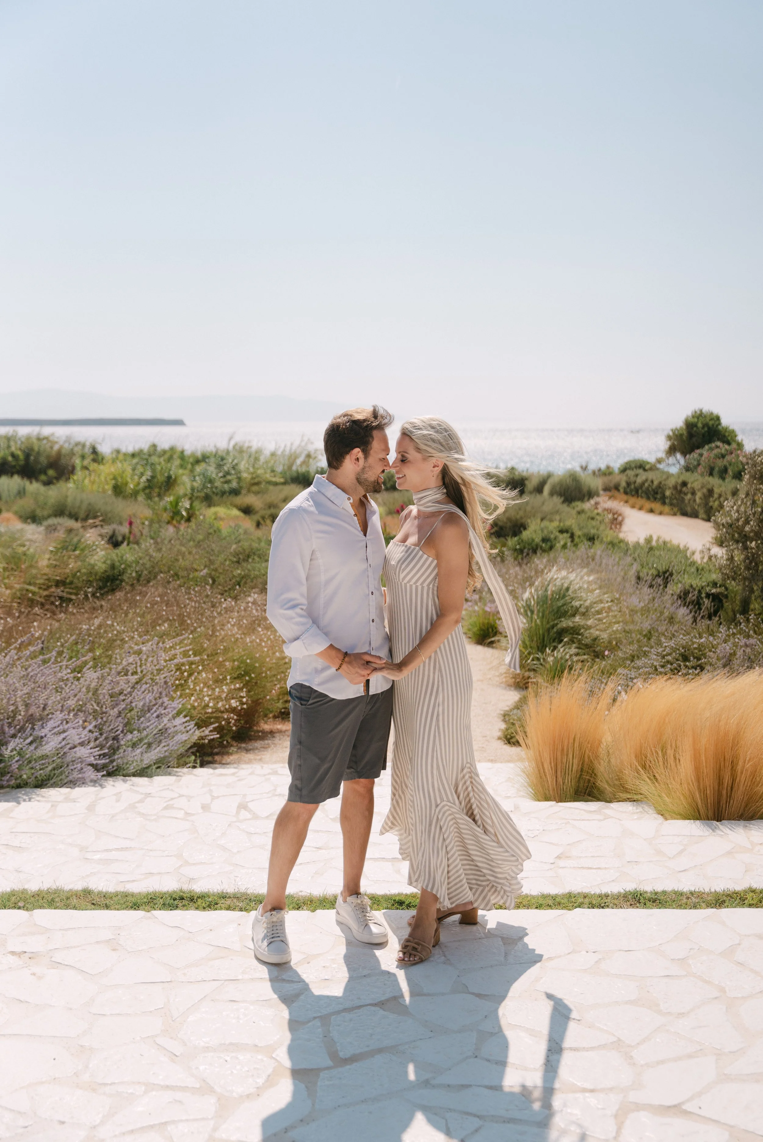 A couple stands close together on a stone pathway by the water, holding hands and smiling at each other, with a lush garden and the ocean in the background.