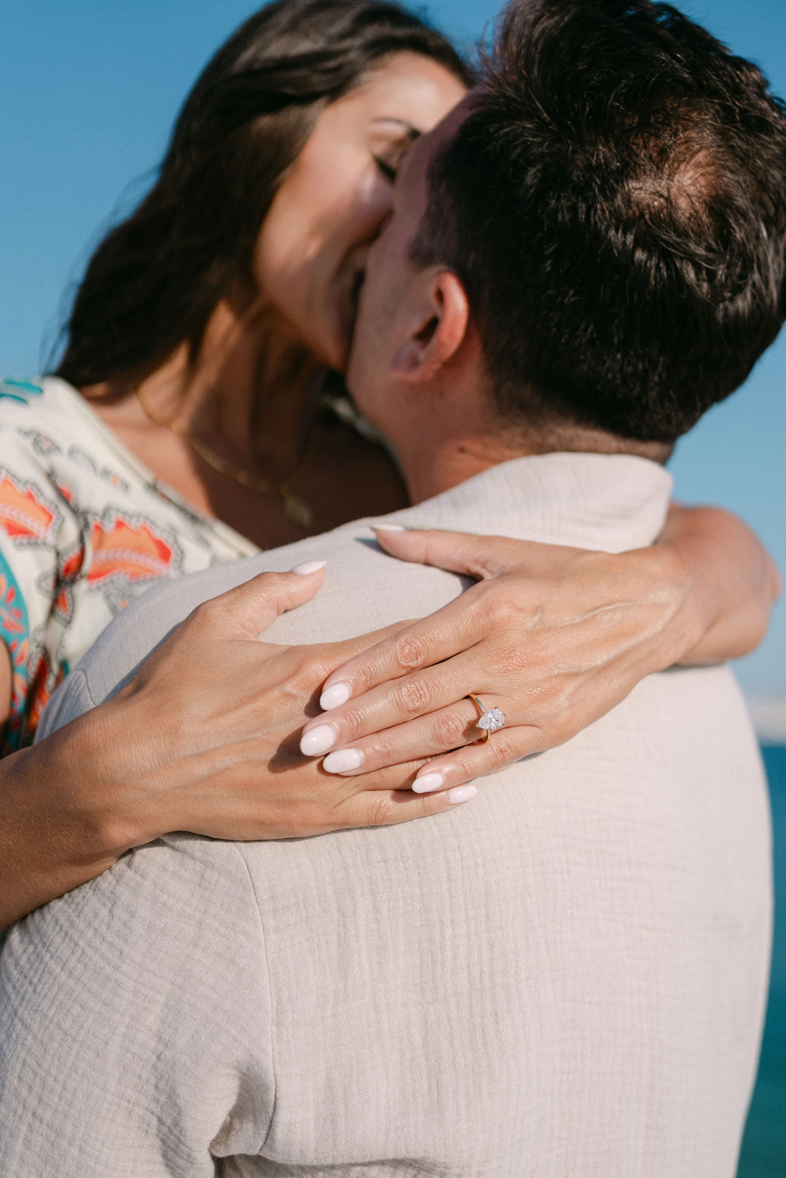 A woman and a man are embracing and kissing on a beach, with the woman's hand showing an engagement ring.