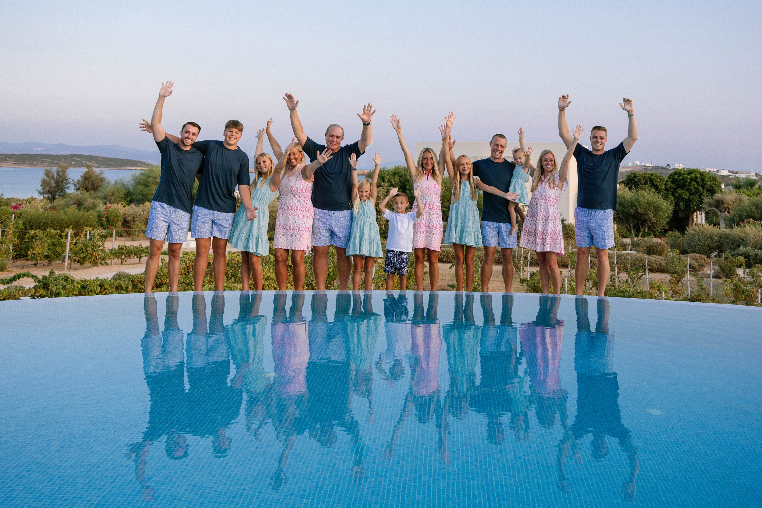 A large family standing by a pool, smiling and waving, with a scenic landscape and water in the background.
