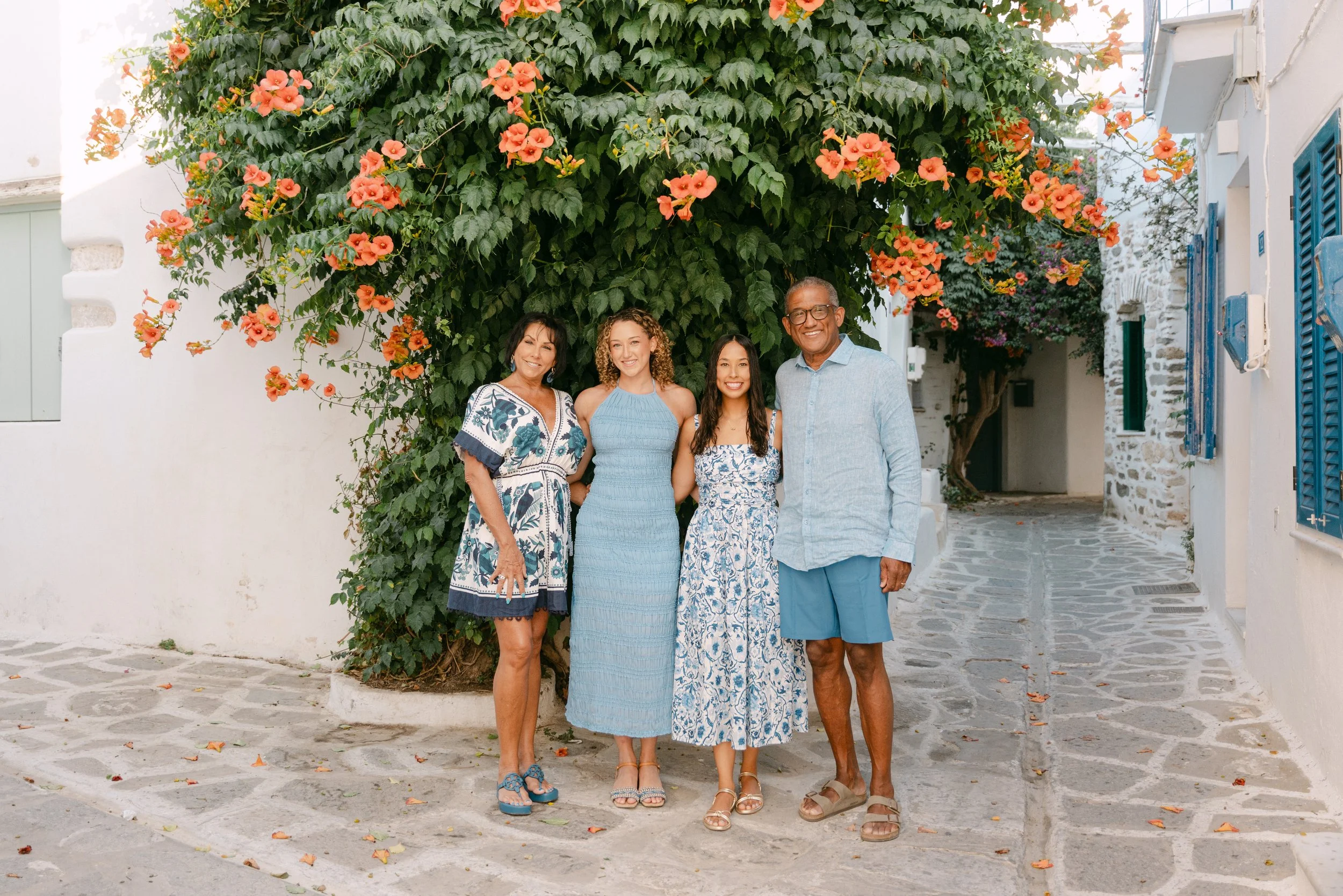 A multigenerational family of five standing on a cobblestone street in front of a flowering orange bougainvillea plant, with white buildings and blue shutters in the background, all dressed in blue and white summer clothes, smiling.