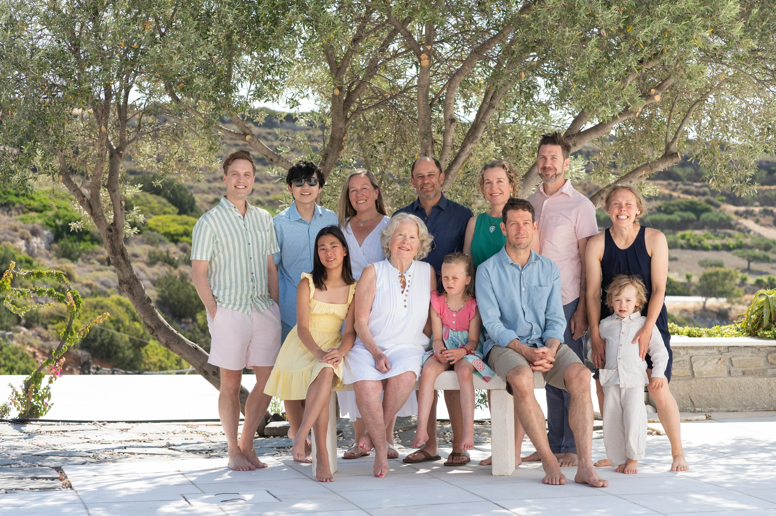 A multigenerational family portrait outdoors with a large tree and green hillside in the background. The family members are smiling and sitting or standing together on a patio, dressed in casual summer clothing.