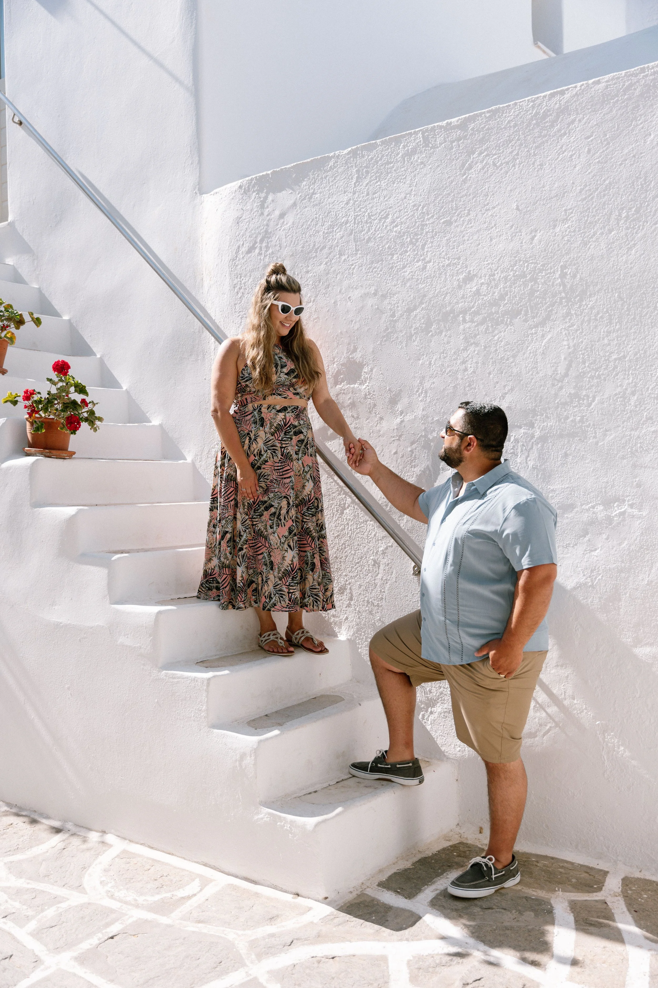 A man on a lower step holding a woman's hand, standing on a staircase in front of a white wall, with potted plants on the stairs, bright sunlight illuminating the scene.