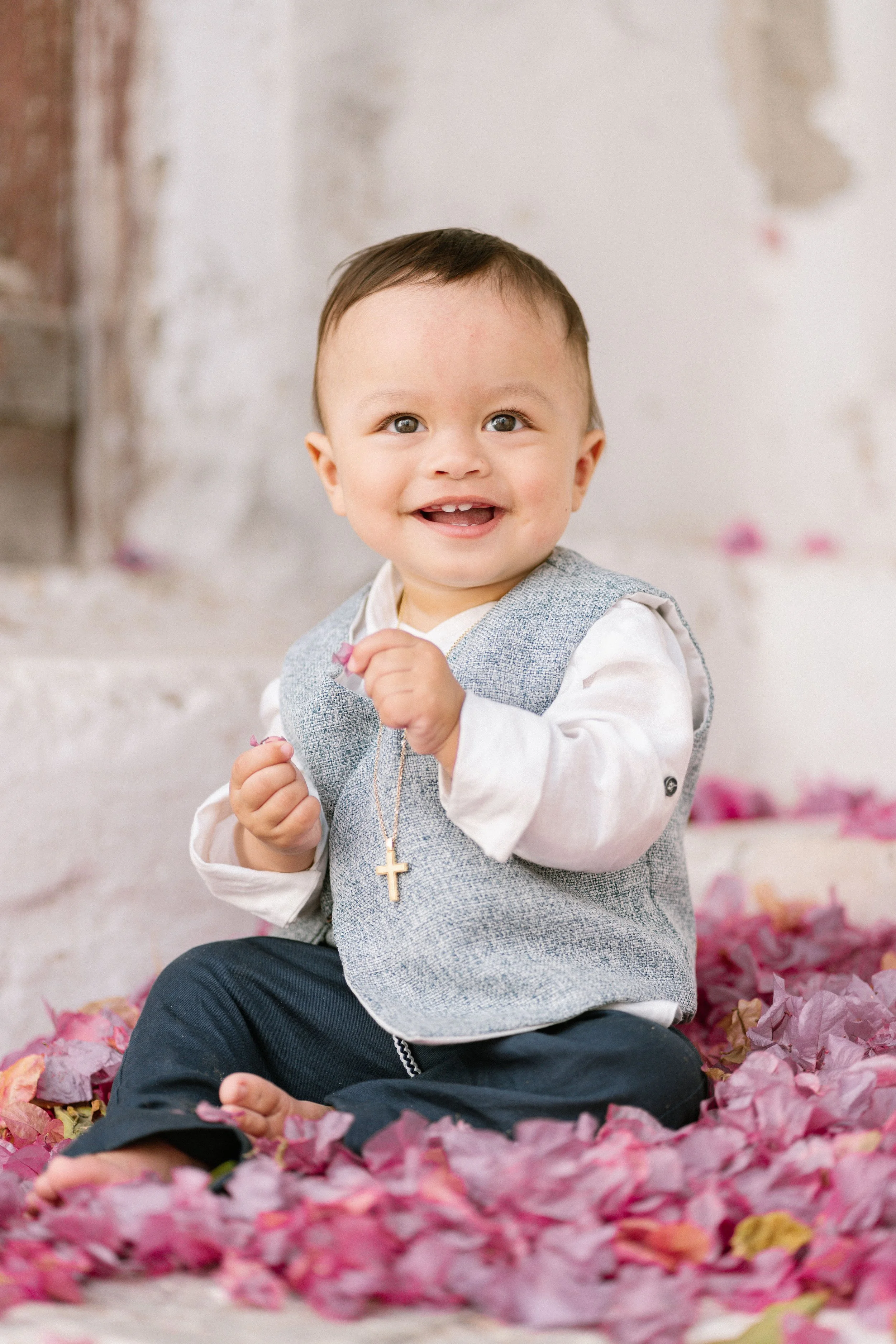 Smiling young child with a gold cross necklace, seated on pink flower petals in front of a white brick wall.