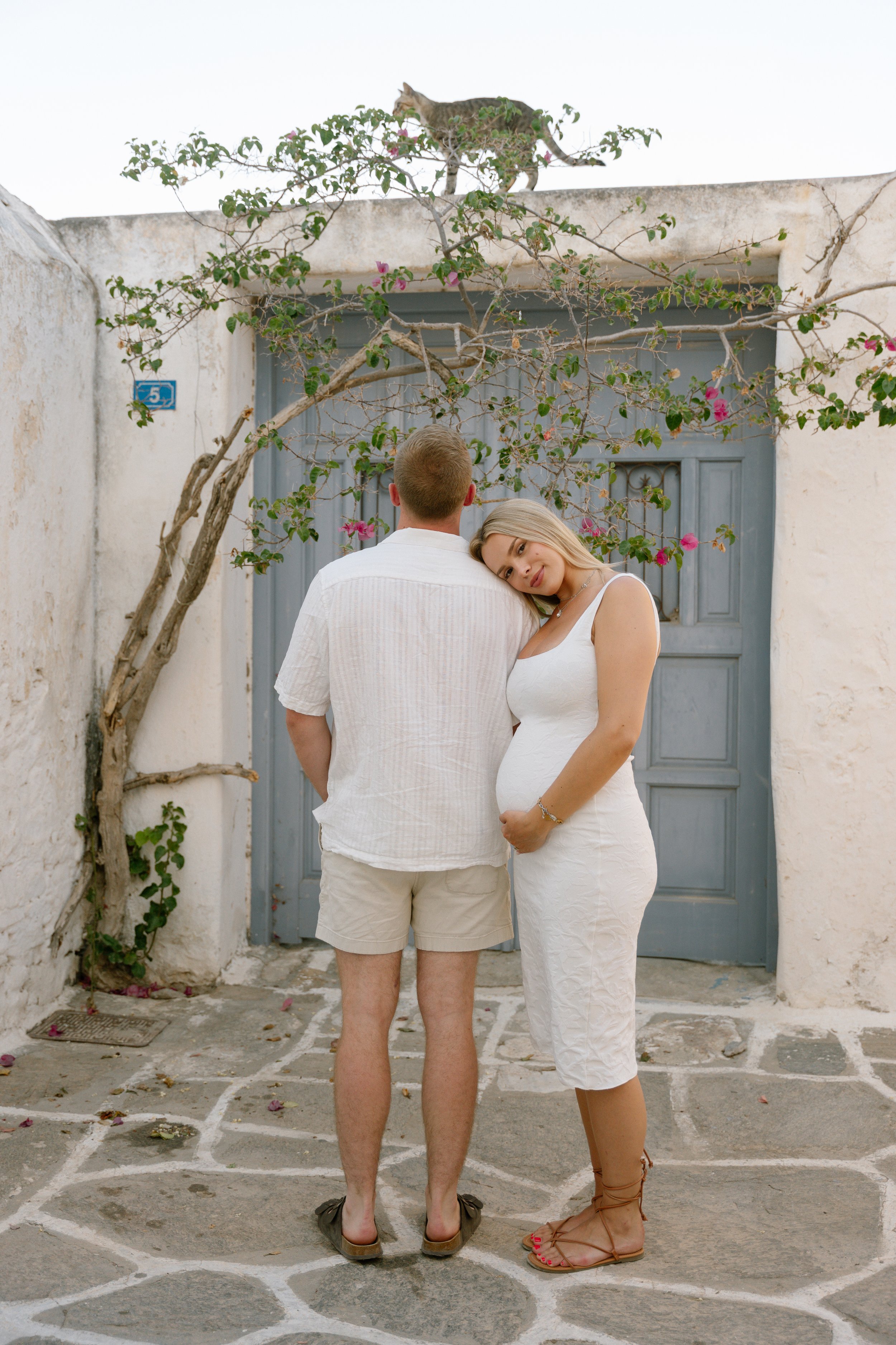 A pregnant woman with blonde hair in a white dress standing next to a man in a white shirt and shorts, with their backs to the camera, outside a white building with a blue door and pink flowering plant. A cat is on top of the building.