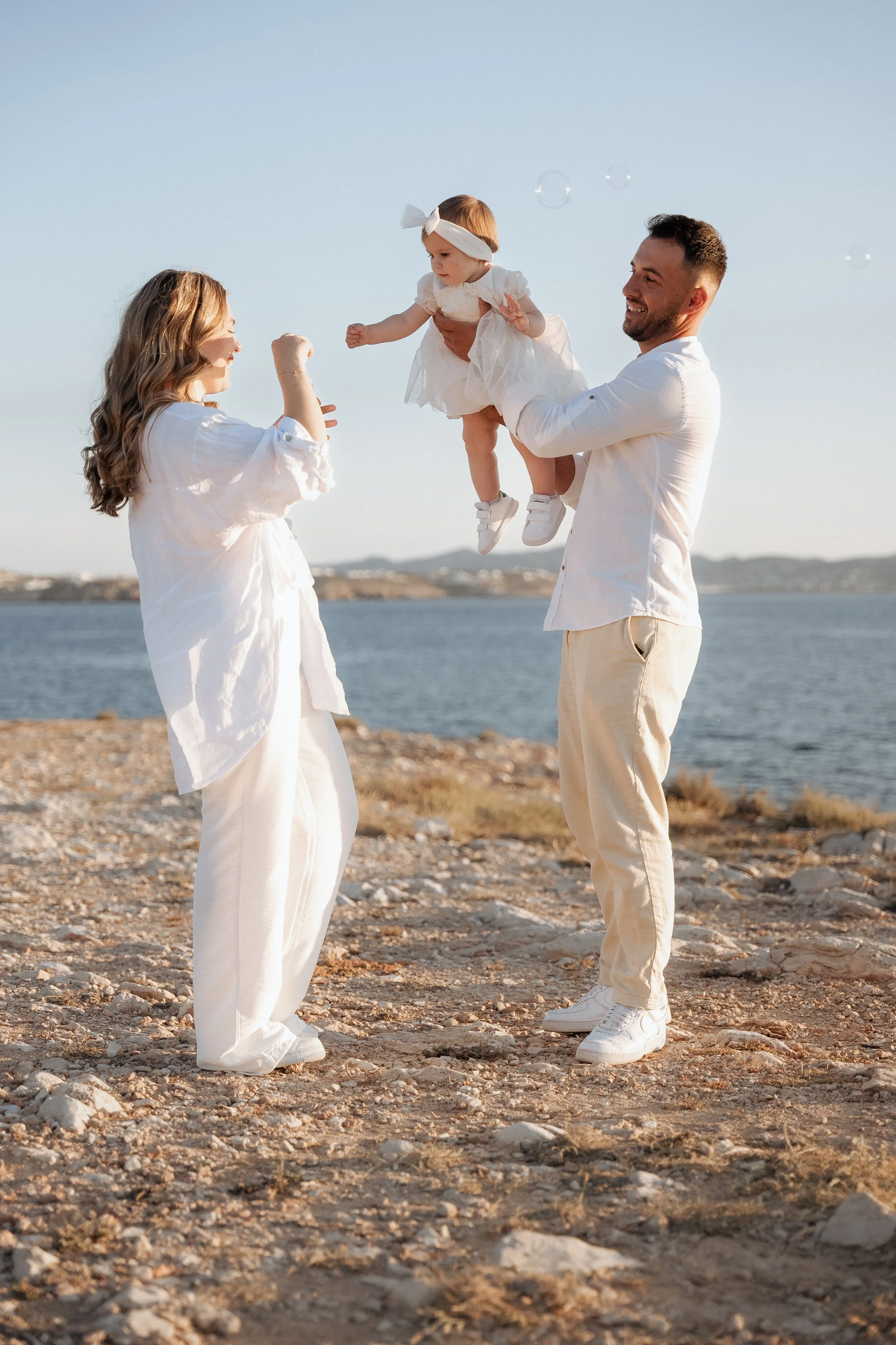 A family of three on a rocky beach by the water, with a woman and man holding a young girl, all dressed in white, playing and smiling together during sunset.