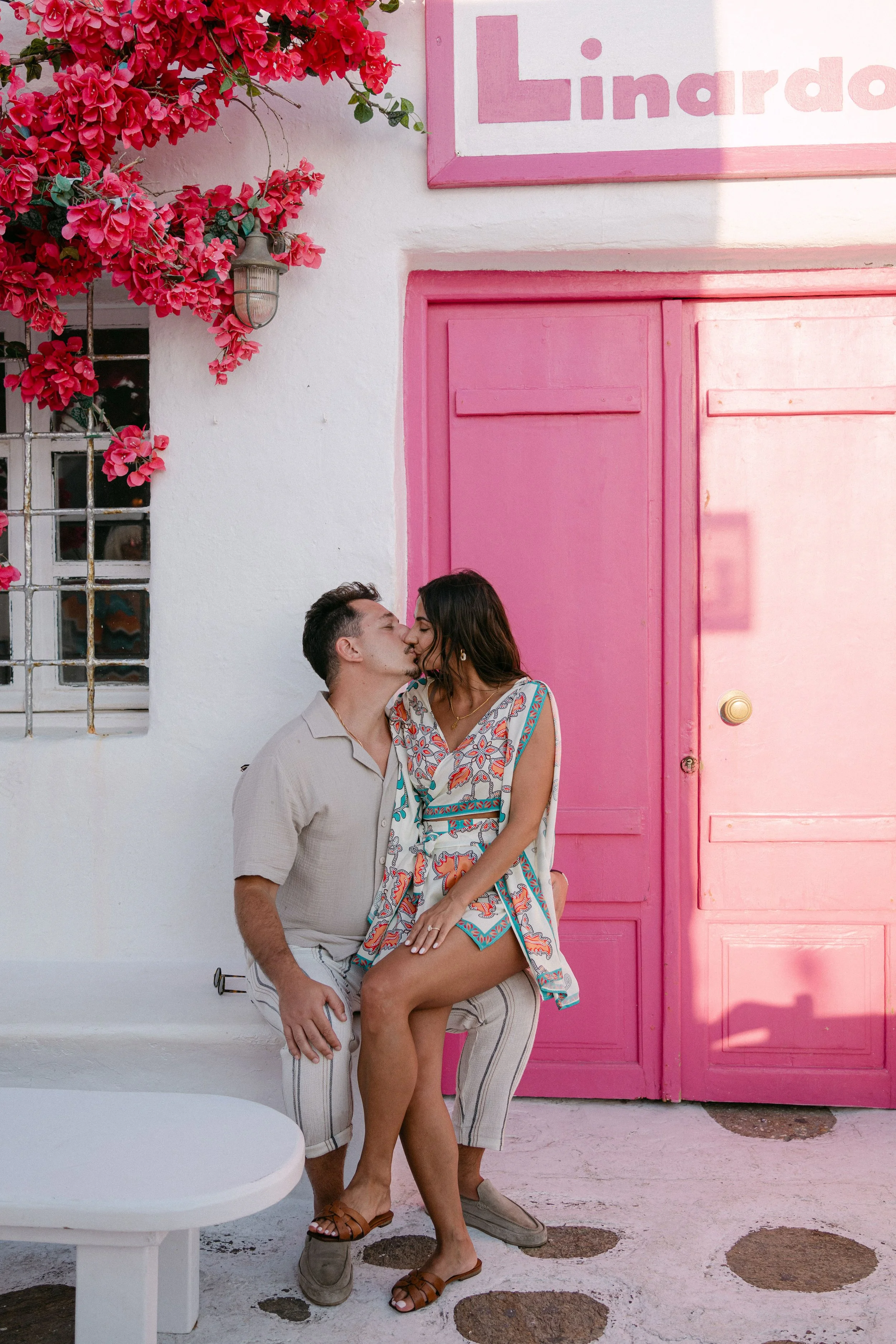 A couple kissing in front of a pink door on a white wall, with pink flowers and a lantern above.