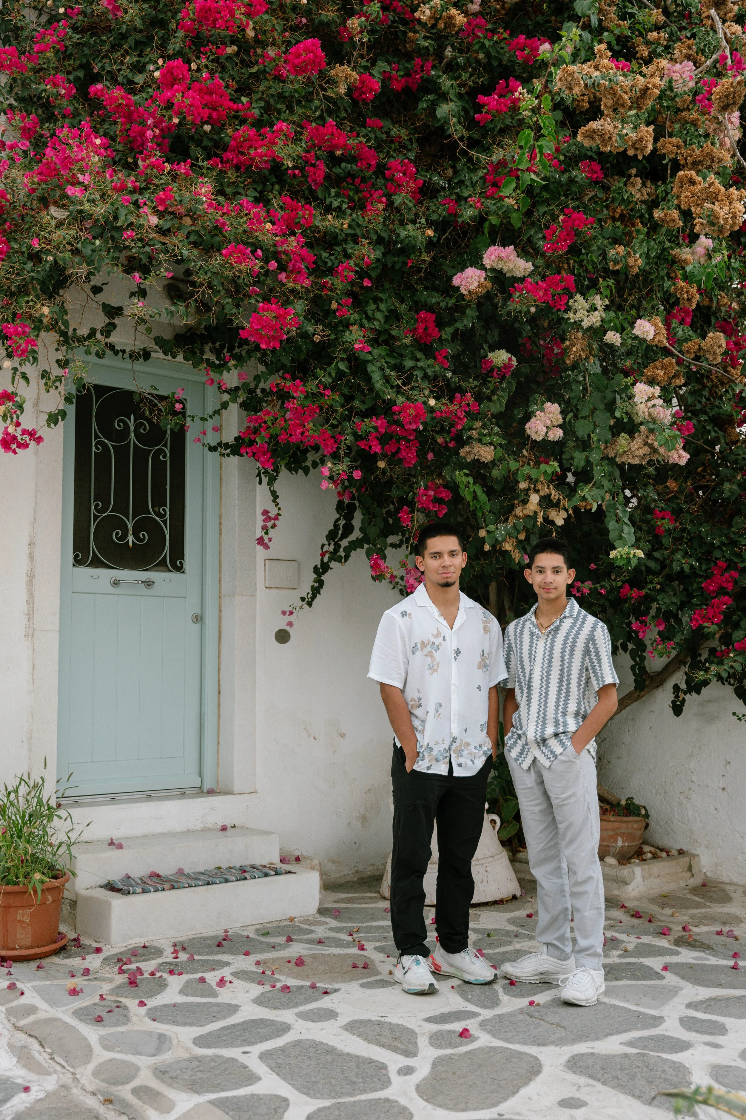 Two young men standing outside in front of a white house with a light blue door and pink bougainvillea flowers overhead.