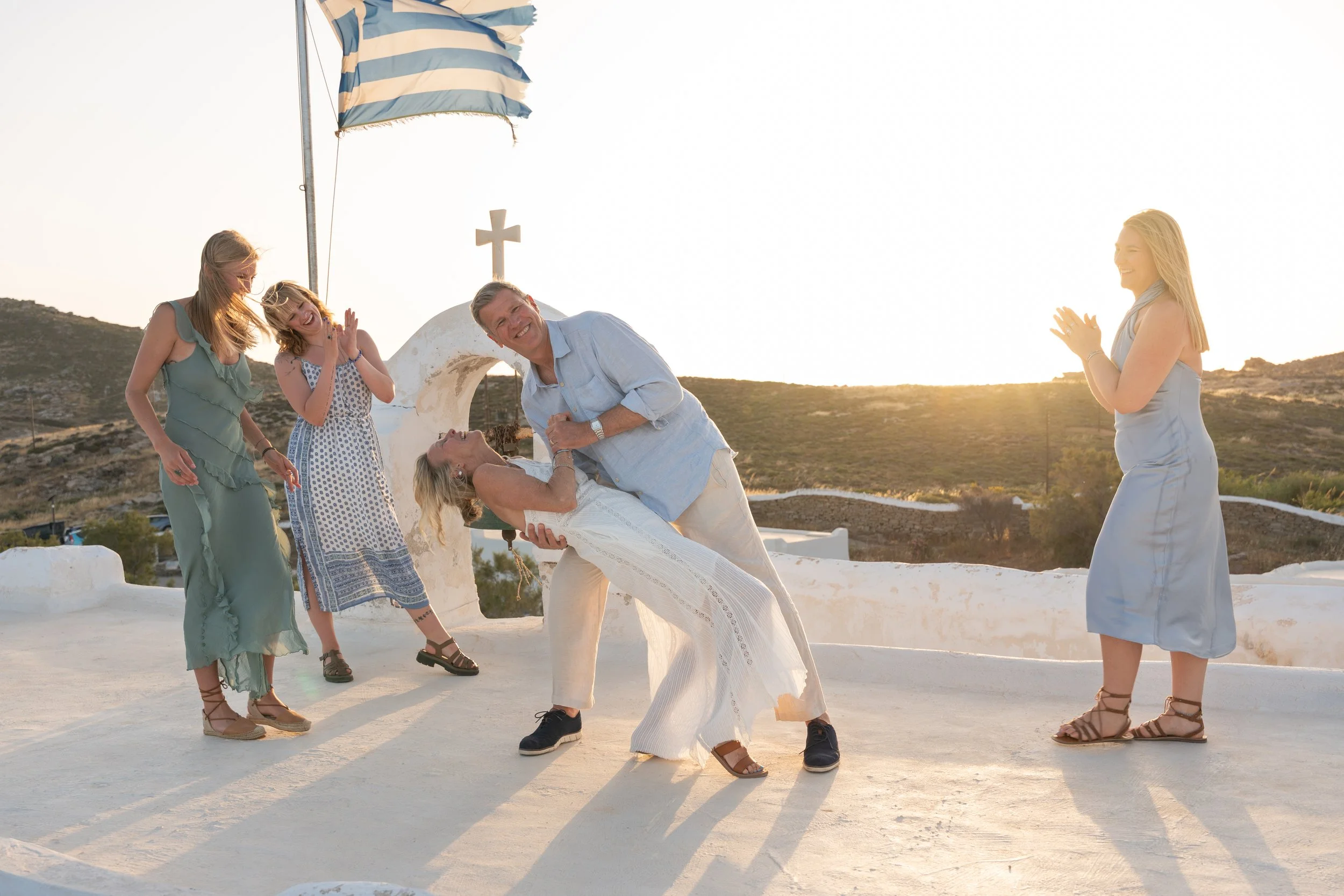 People celebrating a wedding outdoors at sunset, with an ocean view and a white chapel in the background.