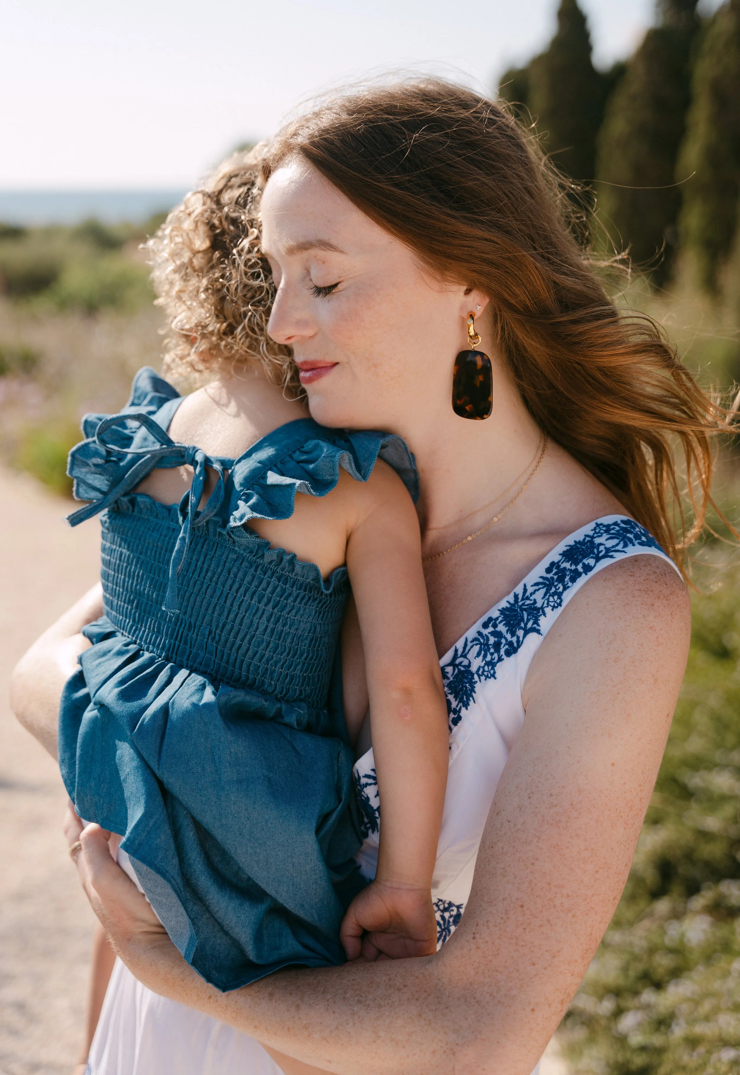 A woman with red hair and freckles hugging a young girl outdoors on a sunny day, with the woman’s eyes closed and a peaceful expression.