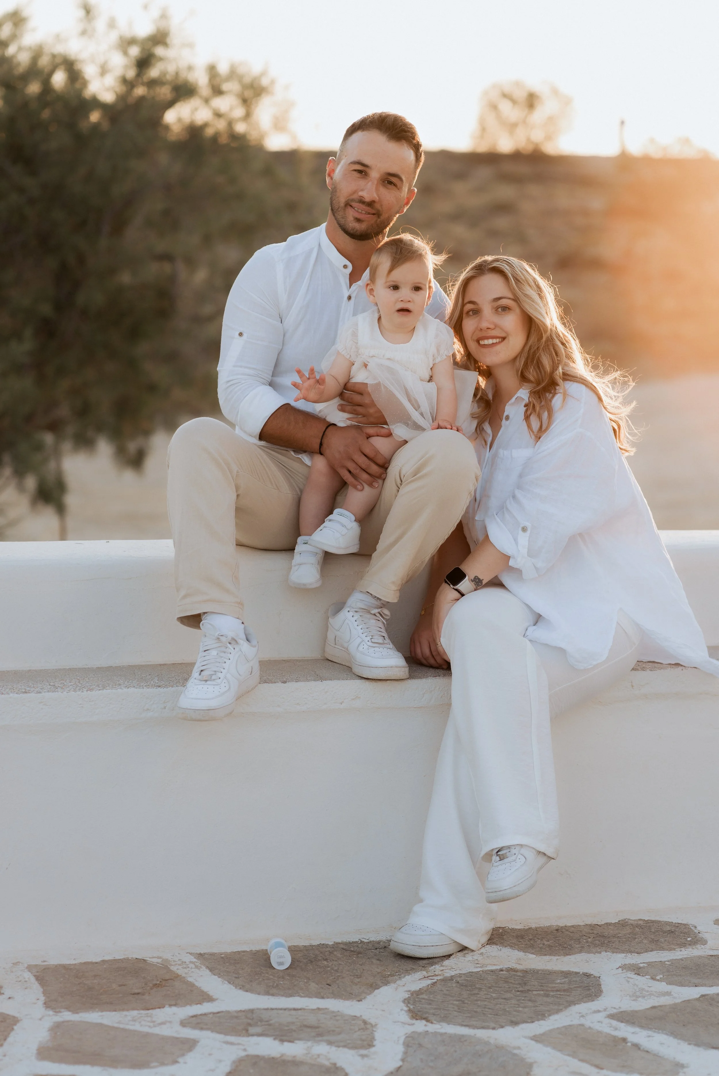 A family of three sitting on a white ledge outdoors during sunset, with trees and a hill in the background. The father, mother, and a young girl are dressed in white and cream-colored clothing, smiling at the camera.