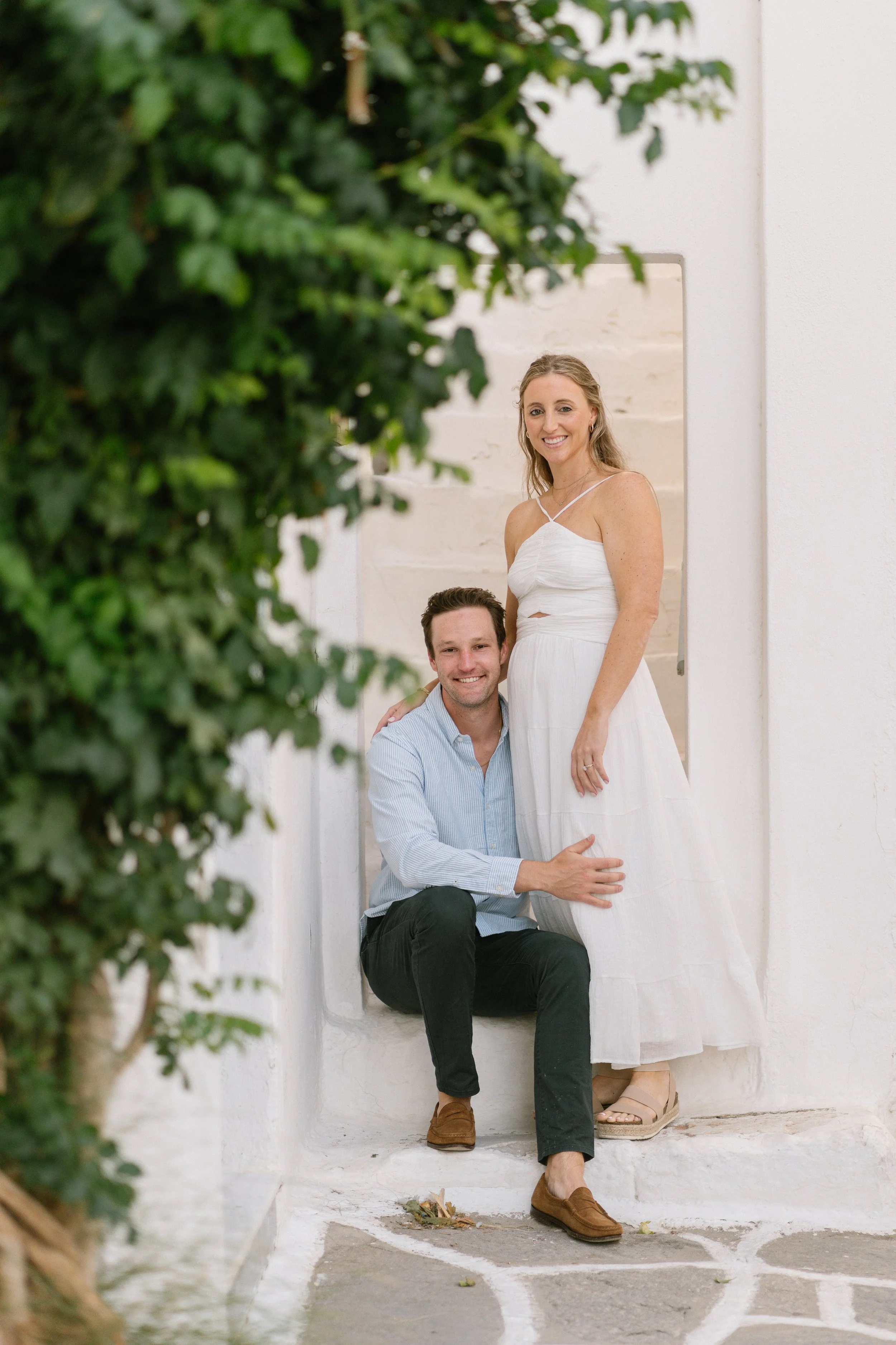 A man and woman pose together on white stairs, with the woman in a white dress standing and the man kneeling, smiling, and touching her leg, framed by green leafy plants on the left.