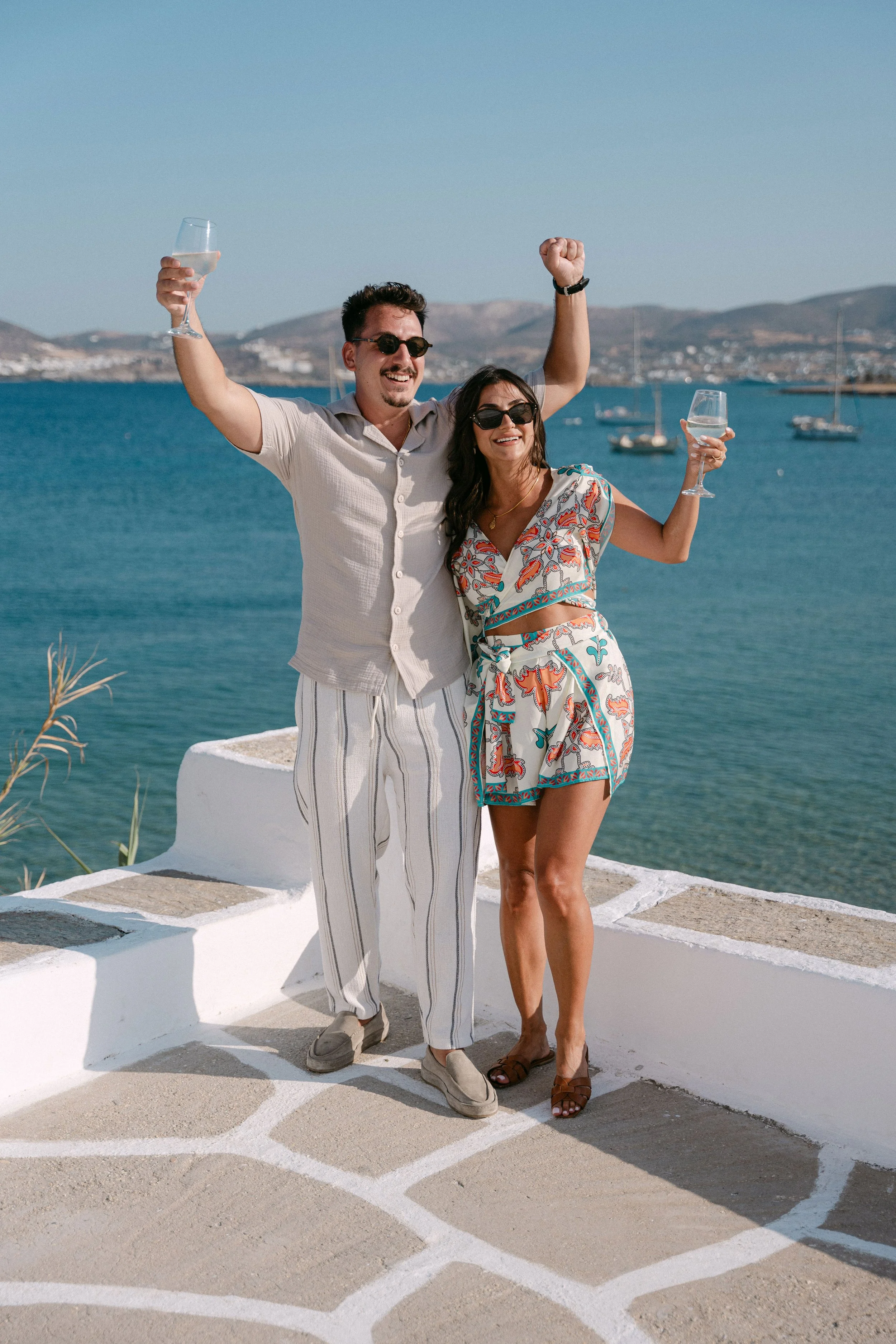 A joyful couple celebrating by the seaside, holding glasses of wine, standing on a white terrace with a view of the ocean and boats in the background.