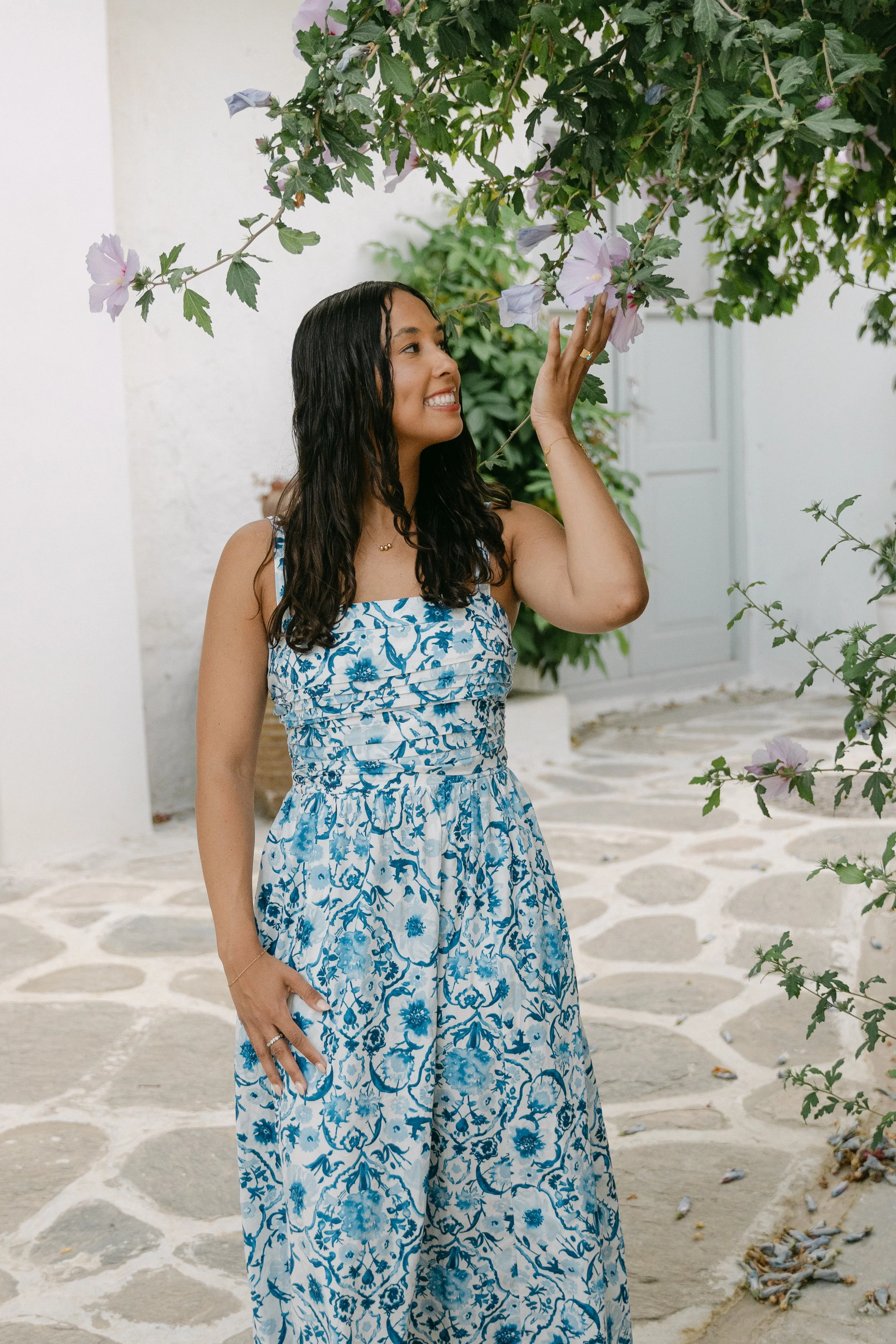 A woman in a blue and white floral dress touches a flowering plant, smiling in a white-walled courtyard with stone paving.