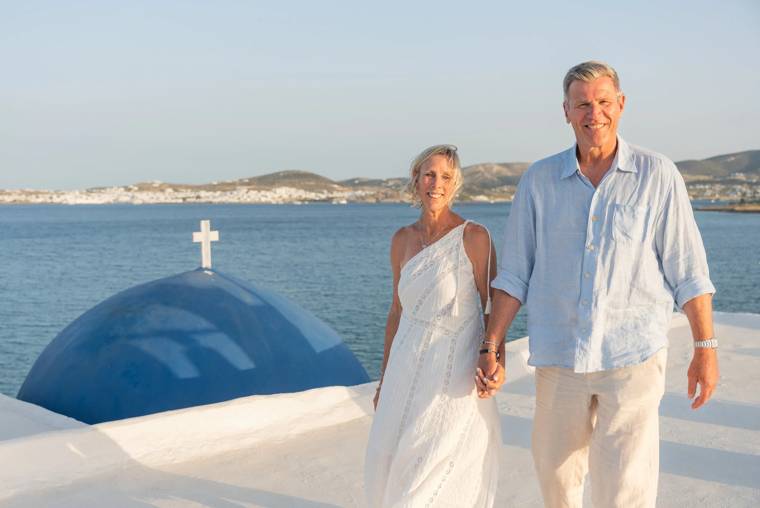 An elderly couple holding hands on a scenic waterfront with a blue-domed church in the background, during sunset or late afternoon, in a Mediterranean setting.