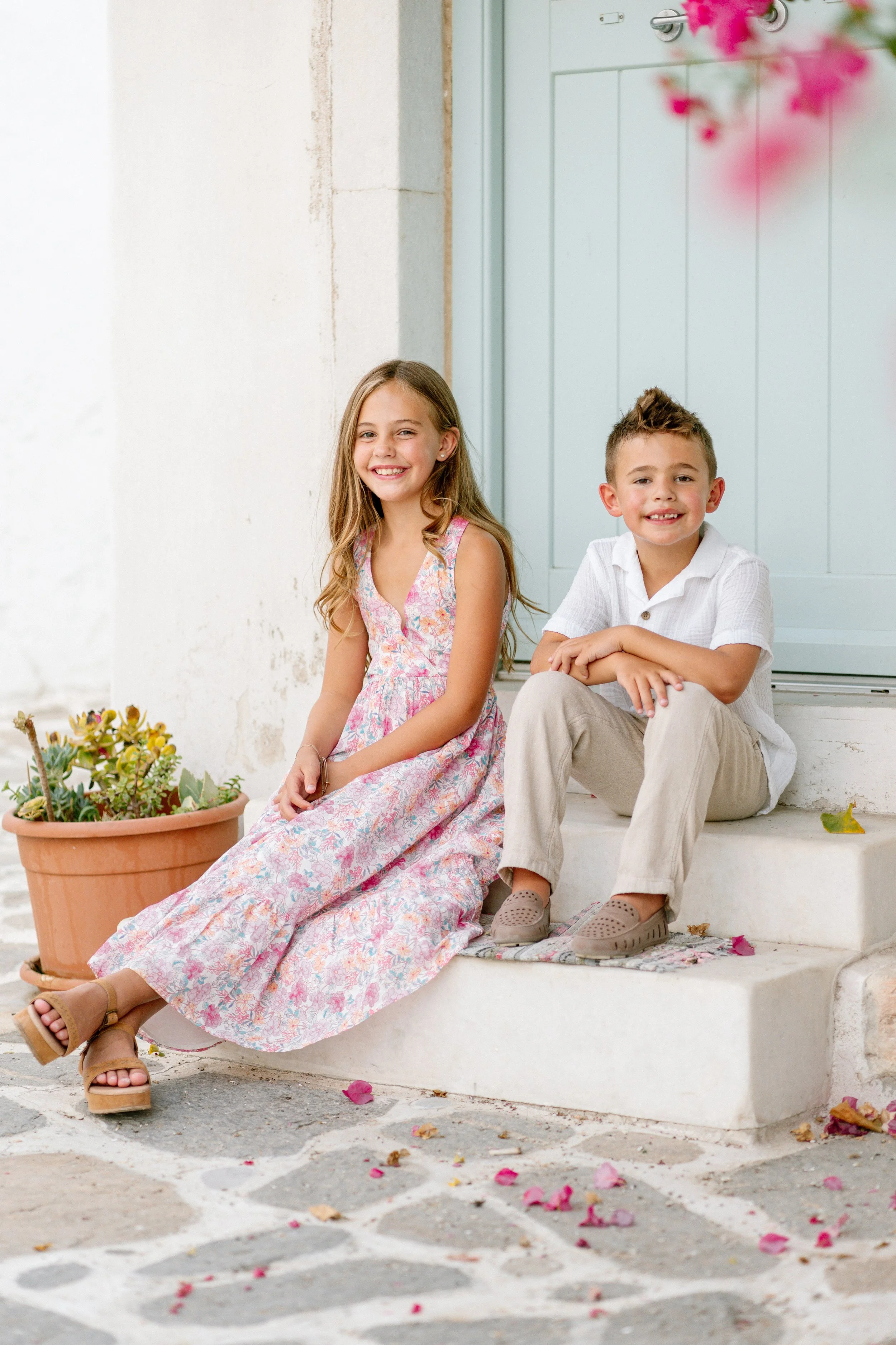 A young girl and boy sitting on front steps of a house, smiling. The girl wears a floral maxi dress and sandals. The boy wears a white shirt, beige pants, and slip-on shoes. There is a potted plant with succulents nearby and fallen flower petals on t