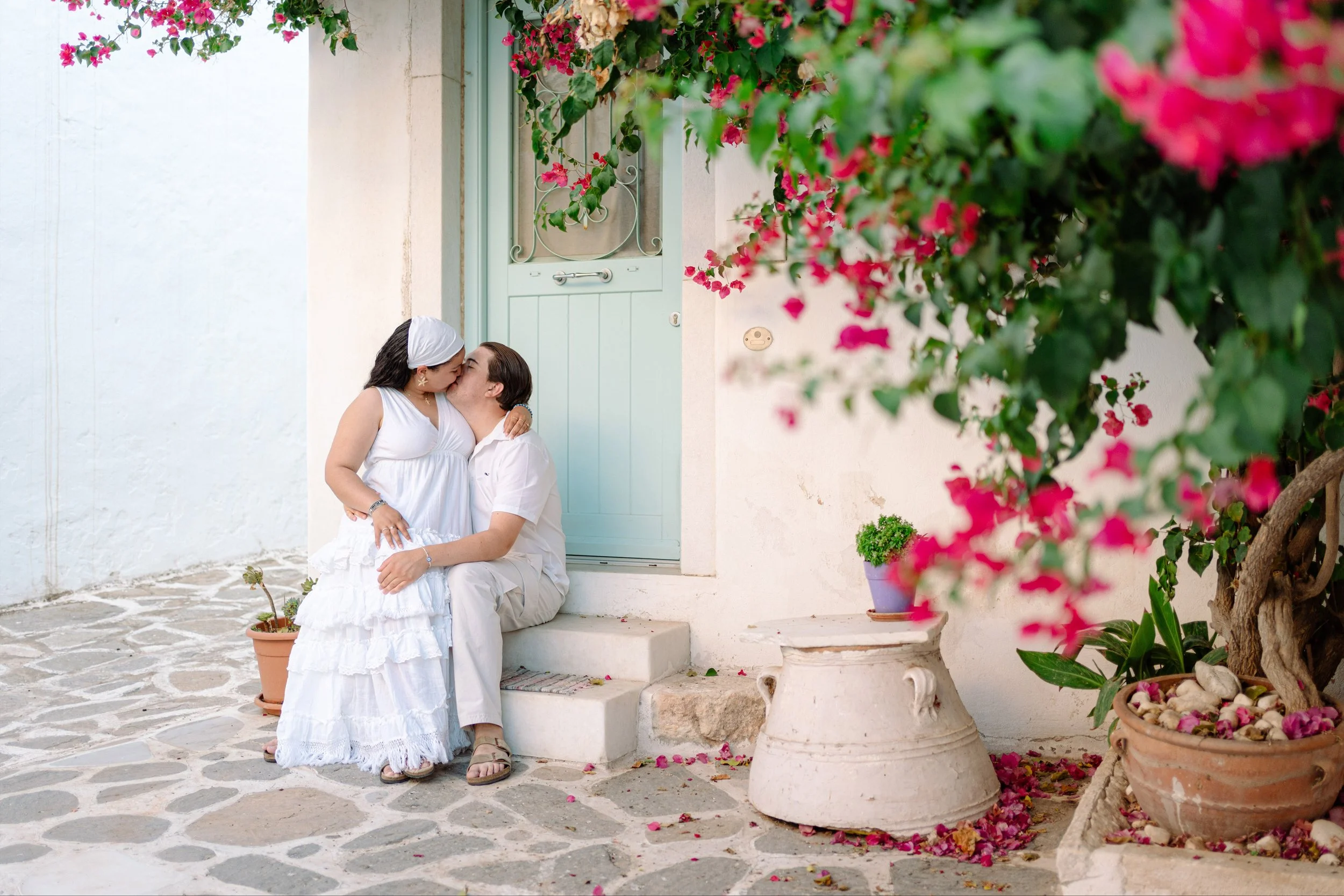 A couple sitting on a step in front of a pastel green door, sharing a kiss. They are dressed in white, with pink bougainvillea flowers hanging above and potted plants on the stone patio.