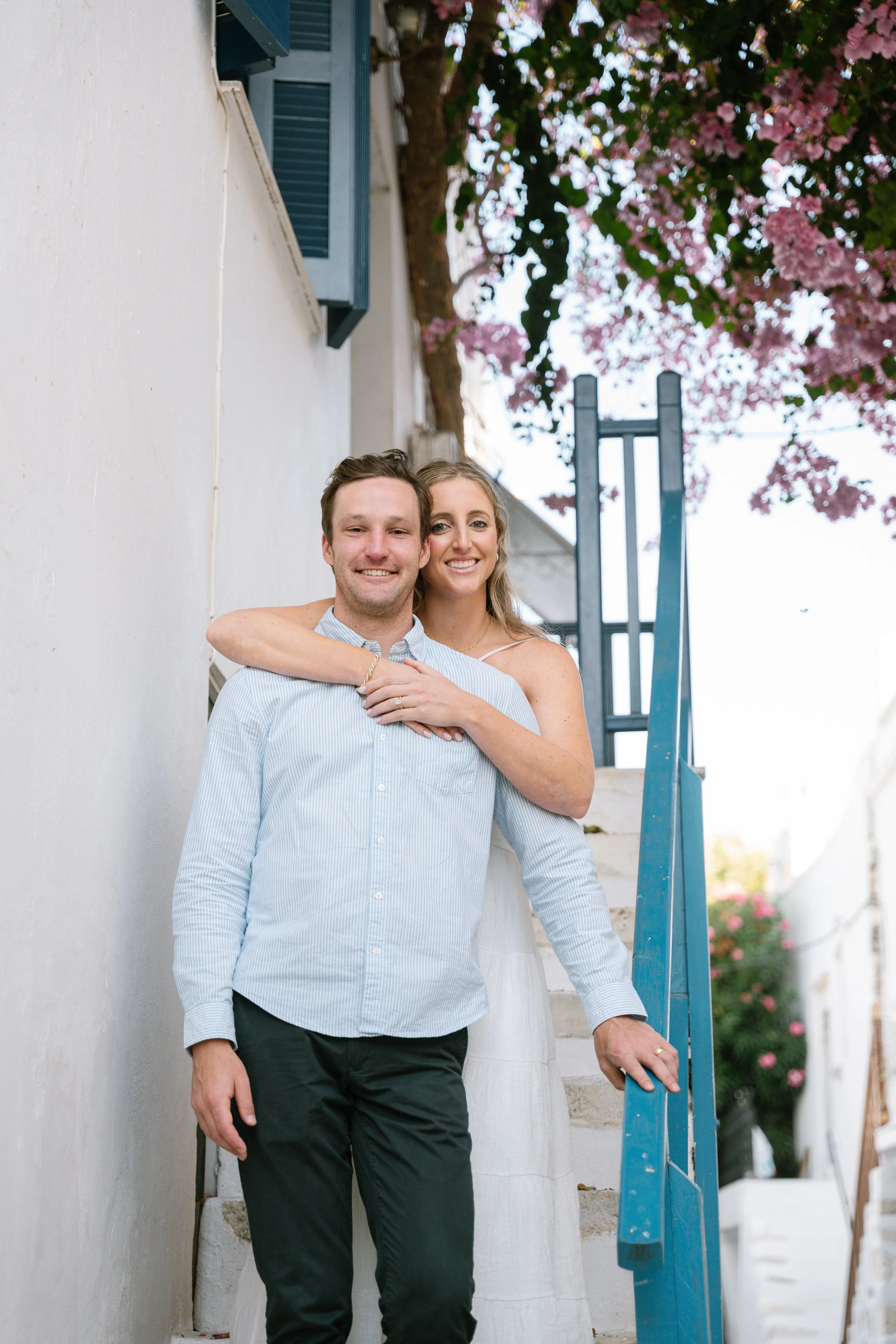 A smiling couple stands on outdoor stairs beside a white wall, with pink blossoms and a blue railing in the background.