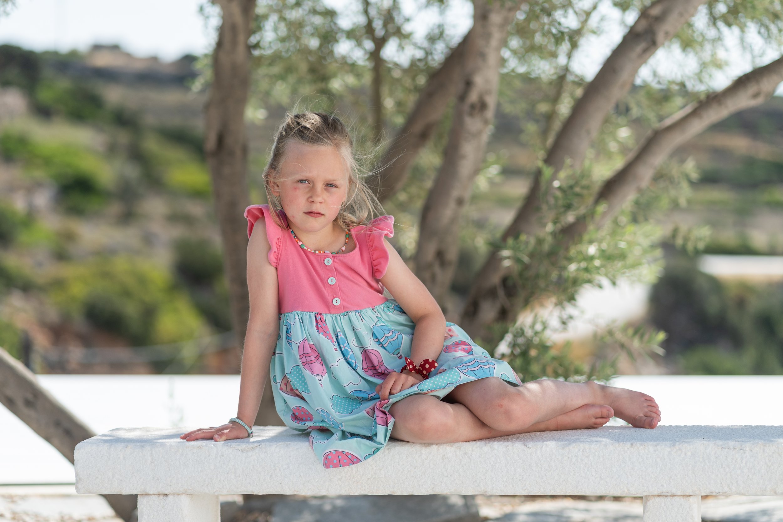 A young girl with blonde hair sitting on a white bench outdoors, wearing a pink and blue dress with a colorful pattern, with trees and a hillside in the background.