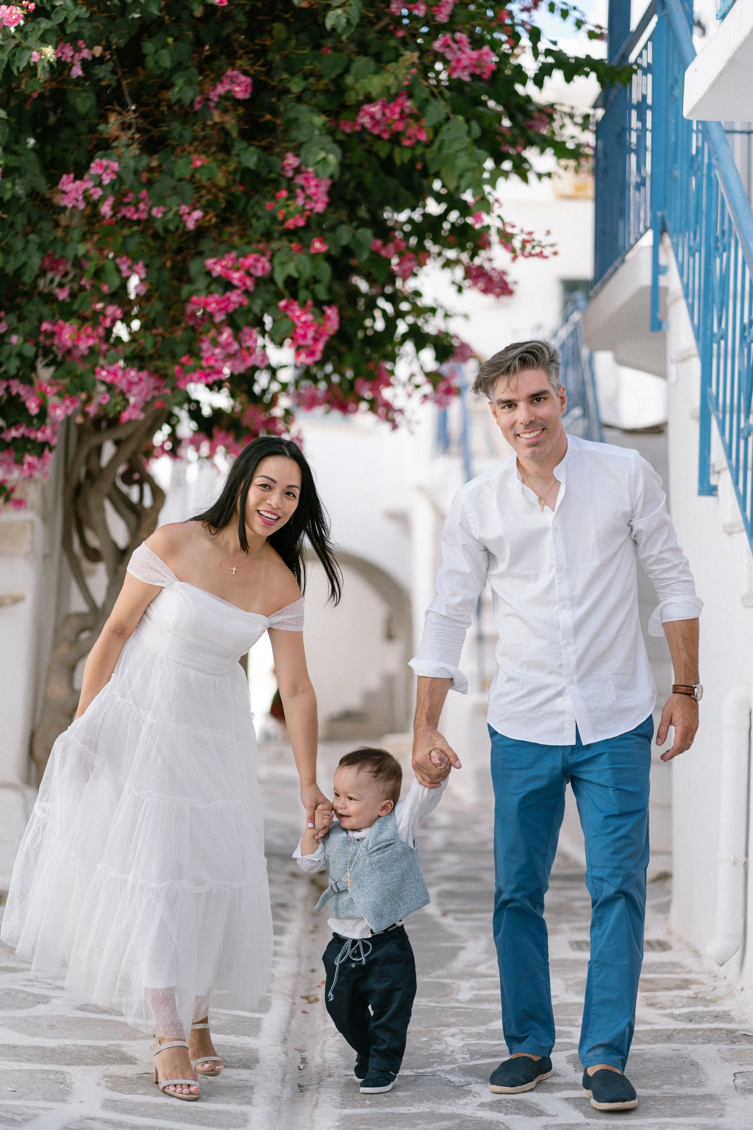 A happy family of three, including a woman, man, and a young boy, walking together hand-in-hand in a picturesque alley with white walls, stairs, and a vibrant pink flowering tree in the background.
