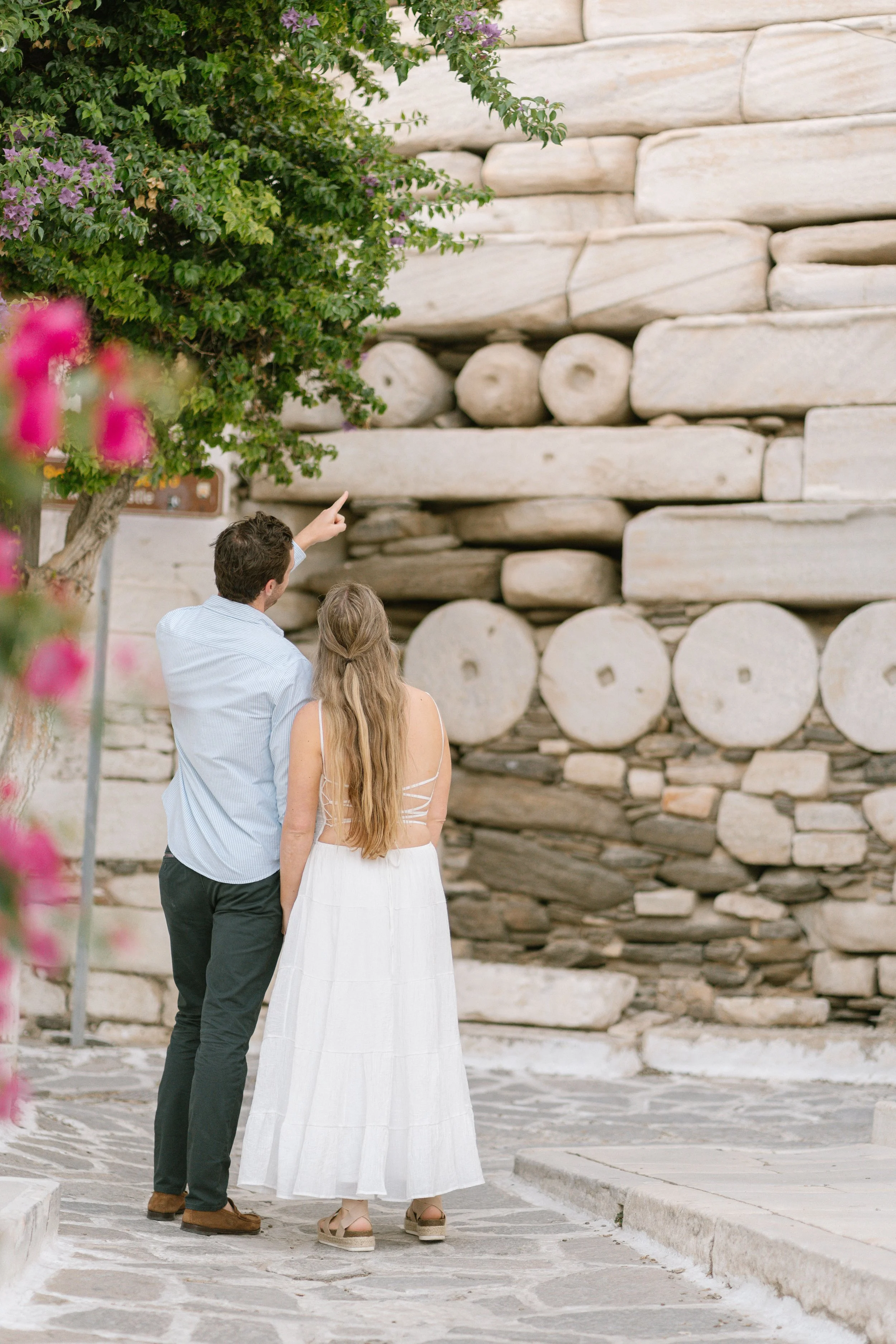 A man and woman in summer clothing stand on stone pavement, looking at a rock wall with logs and stones, with pink flowers and green leaves nearby.