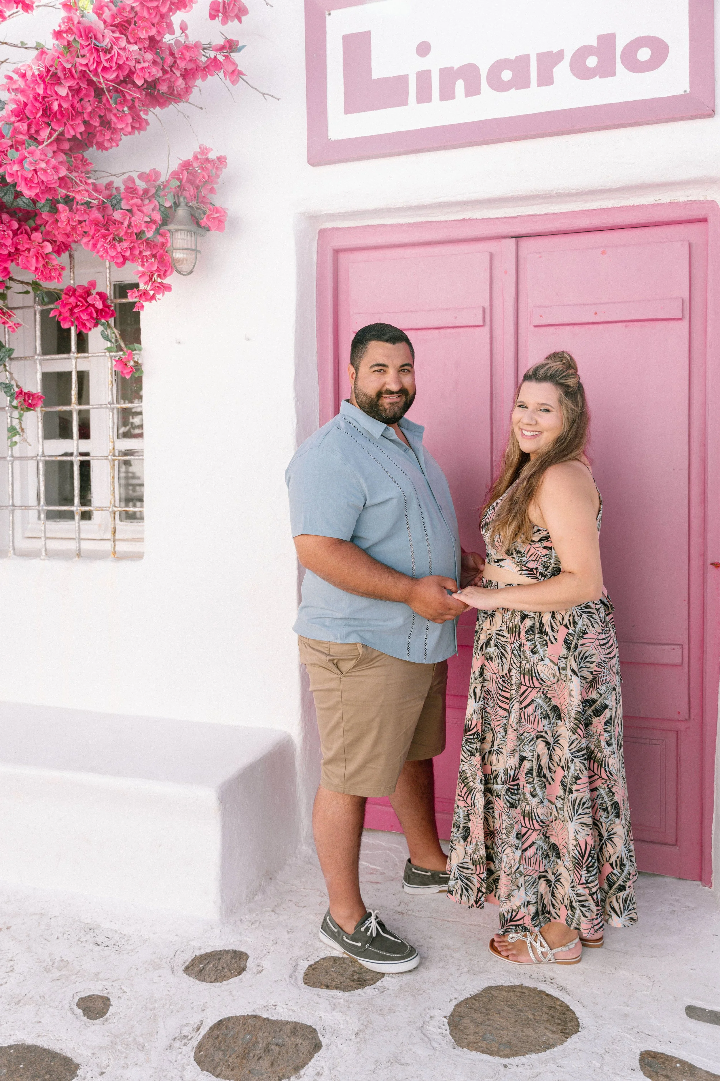 A man and woman holding hands in front of a pink door with a sign that says 'Linado', with pink bougainvillaea flowers on the side.