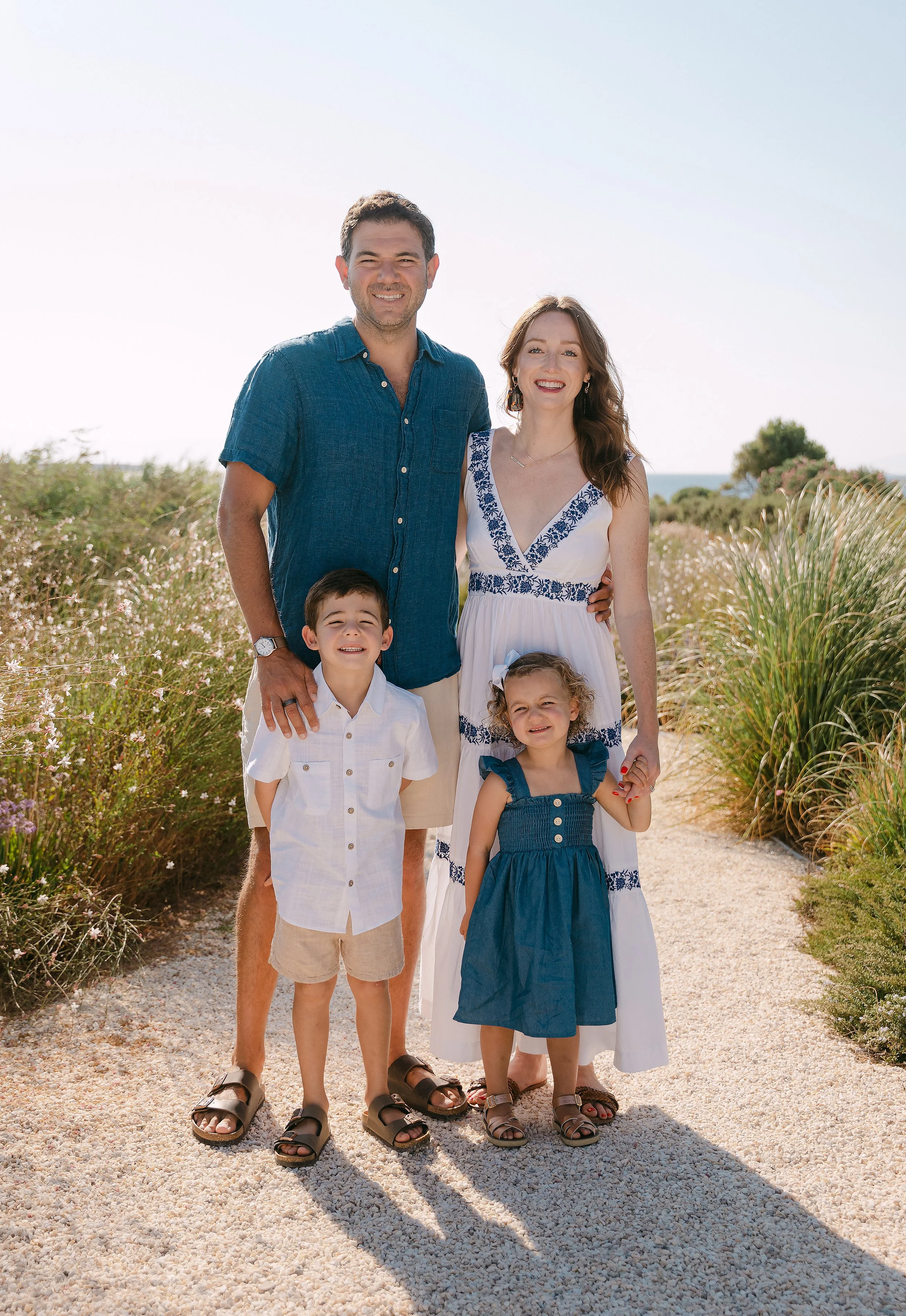 A family of four standing on a sandy path outdoors, with beach and ocean in the background. The father wears a blue shirt, shorts, and sandals. The mother wears a white dress with blue embroidery. The son and daughter wear white and blue dresses, res