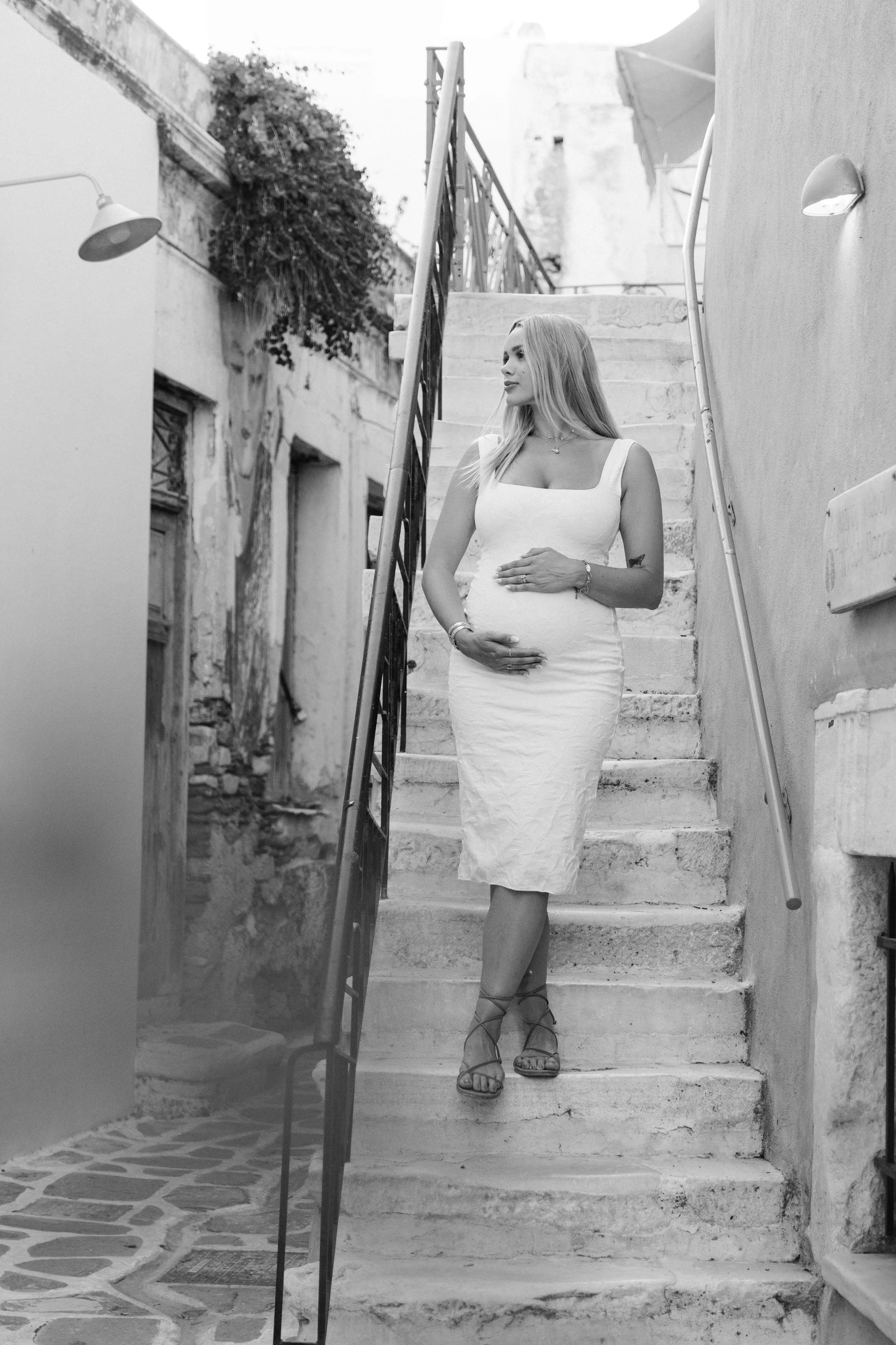 A pregnant woman in a white dress standing on a stone staircase in an outdoor alleyway, touching her belly and looking to the side.