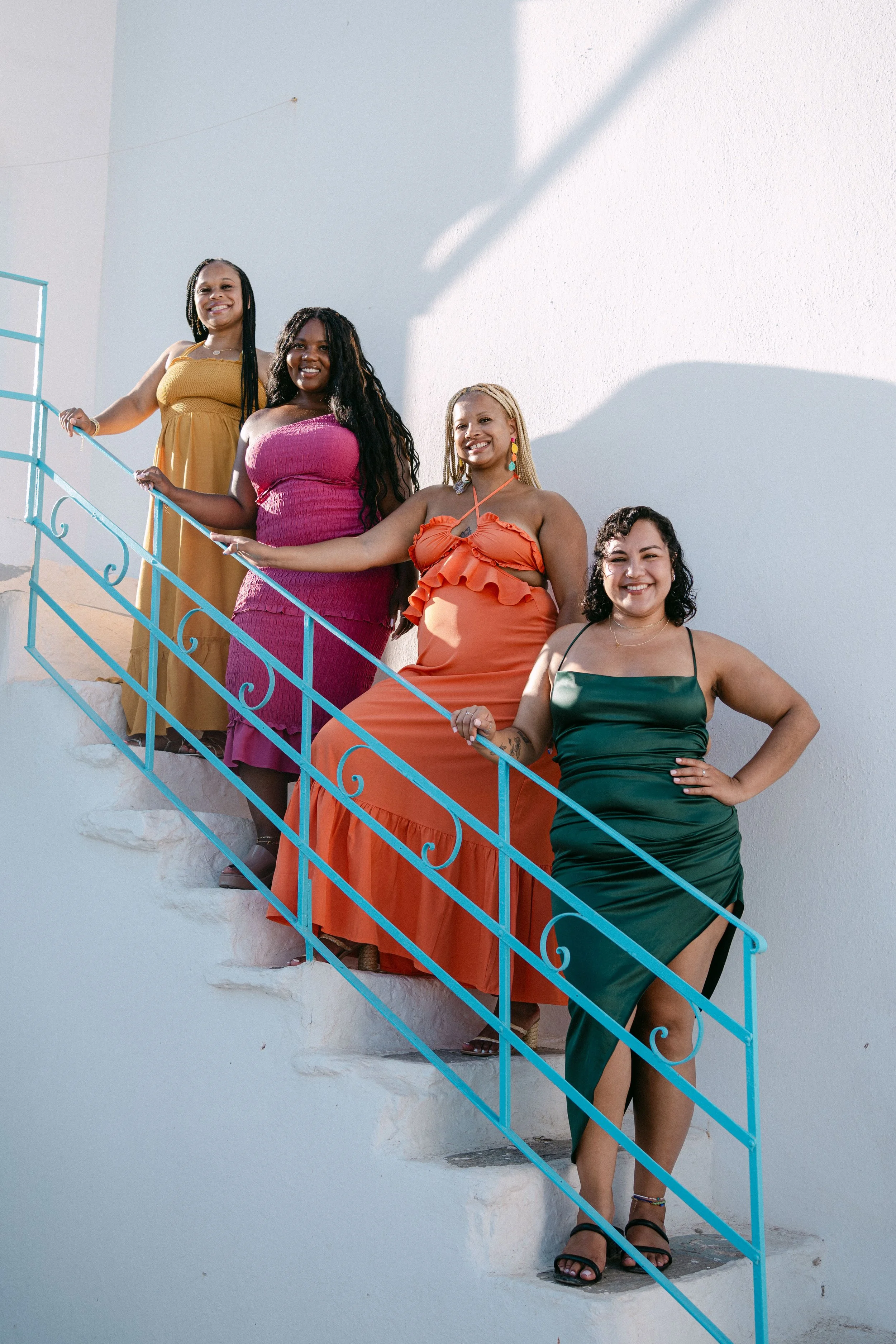 Group of five women standing on a white staircase outside, smiling and dressed in colorful dresses, against a white wall with sunlight and shadows.