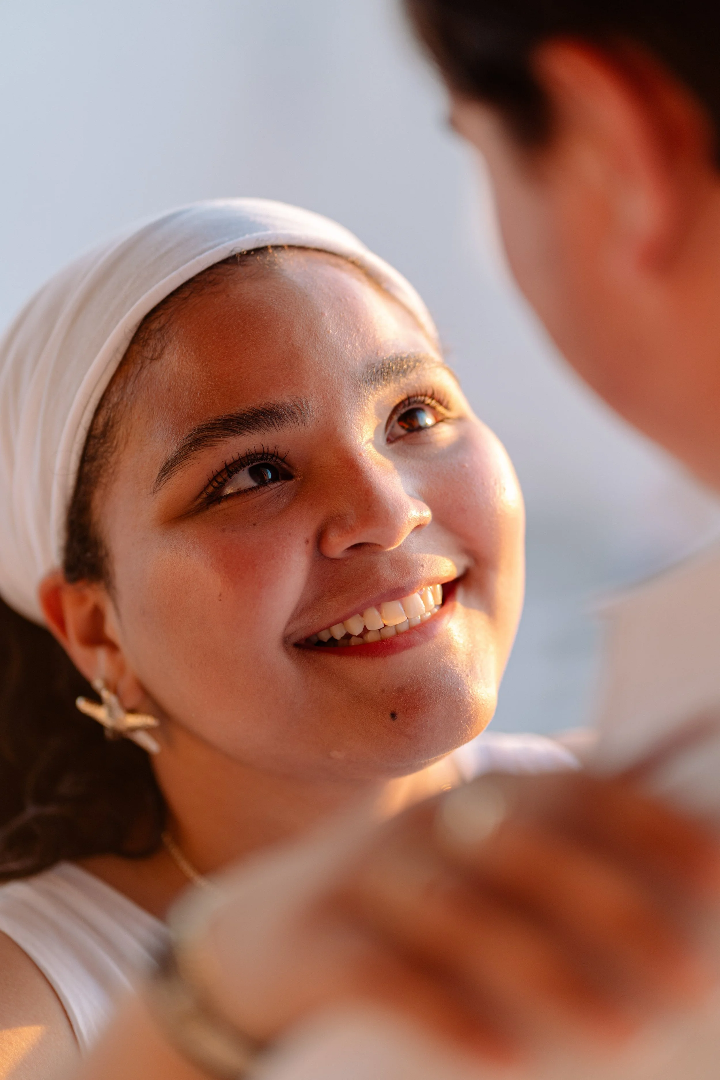 A woman with a white headband and earrings is smiling and looking at someone off-camera during sunset.