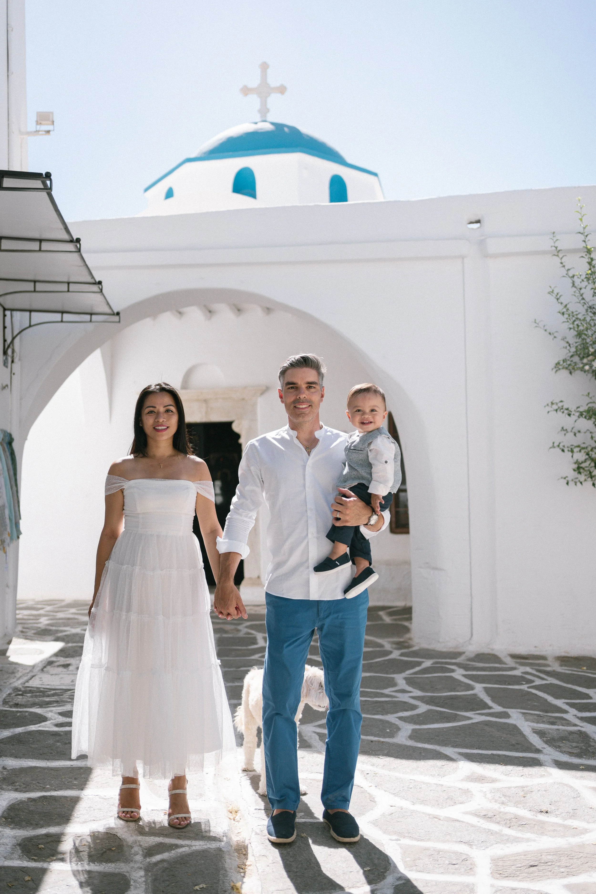 A family of three walking hand-in-hand outside a white building with a blue dome and cross, likely a church, on a sunny day.