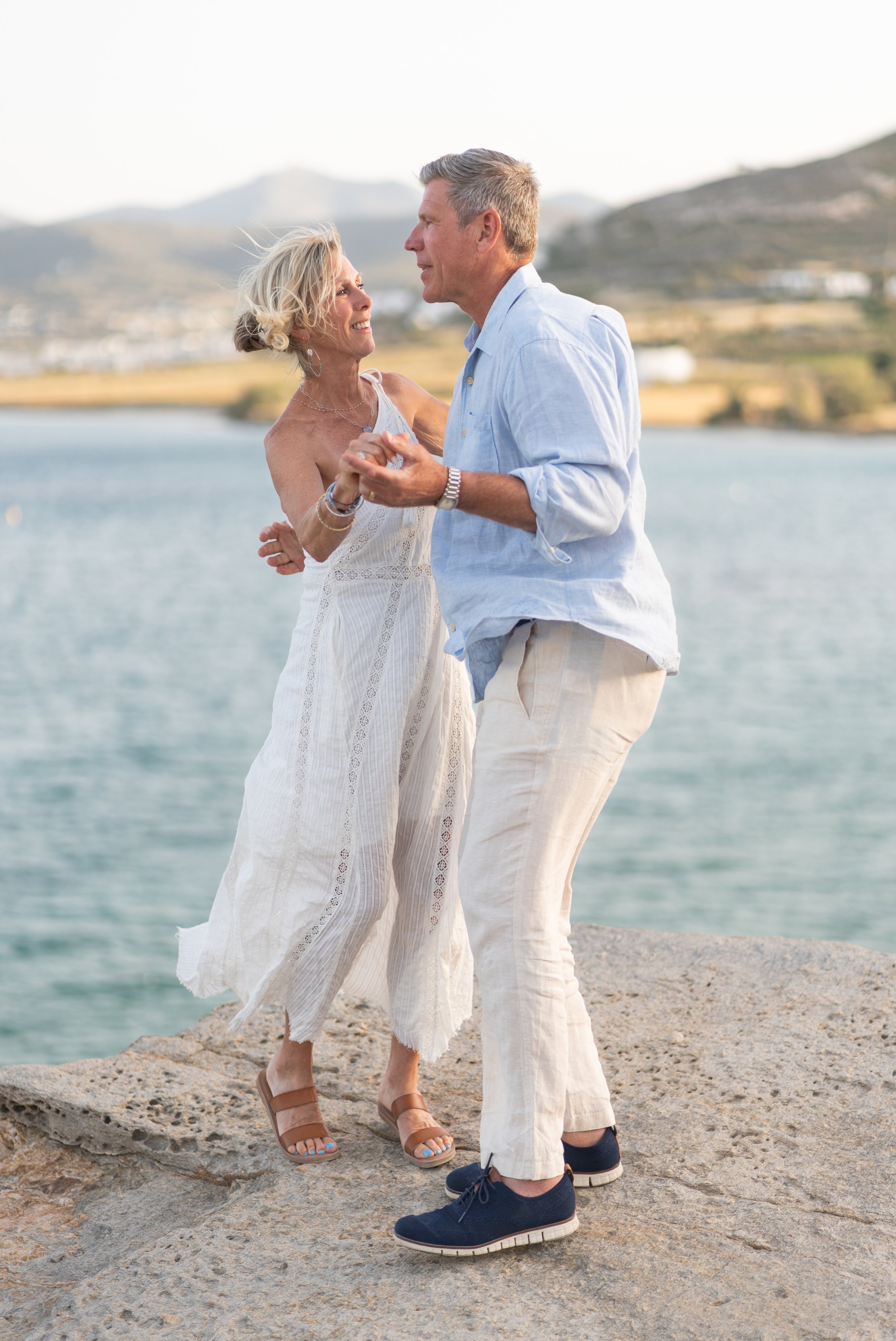 An older couple dancing by the water with a hillside in the background.