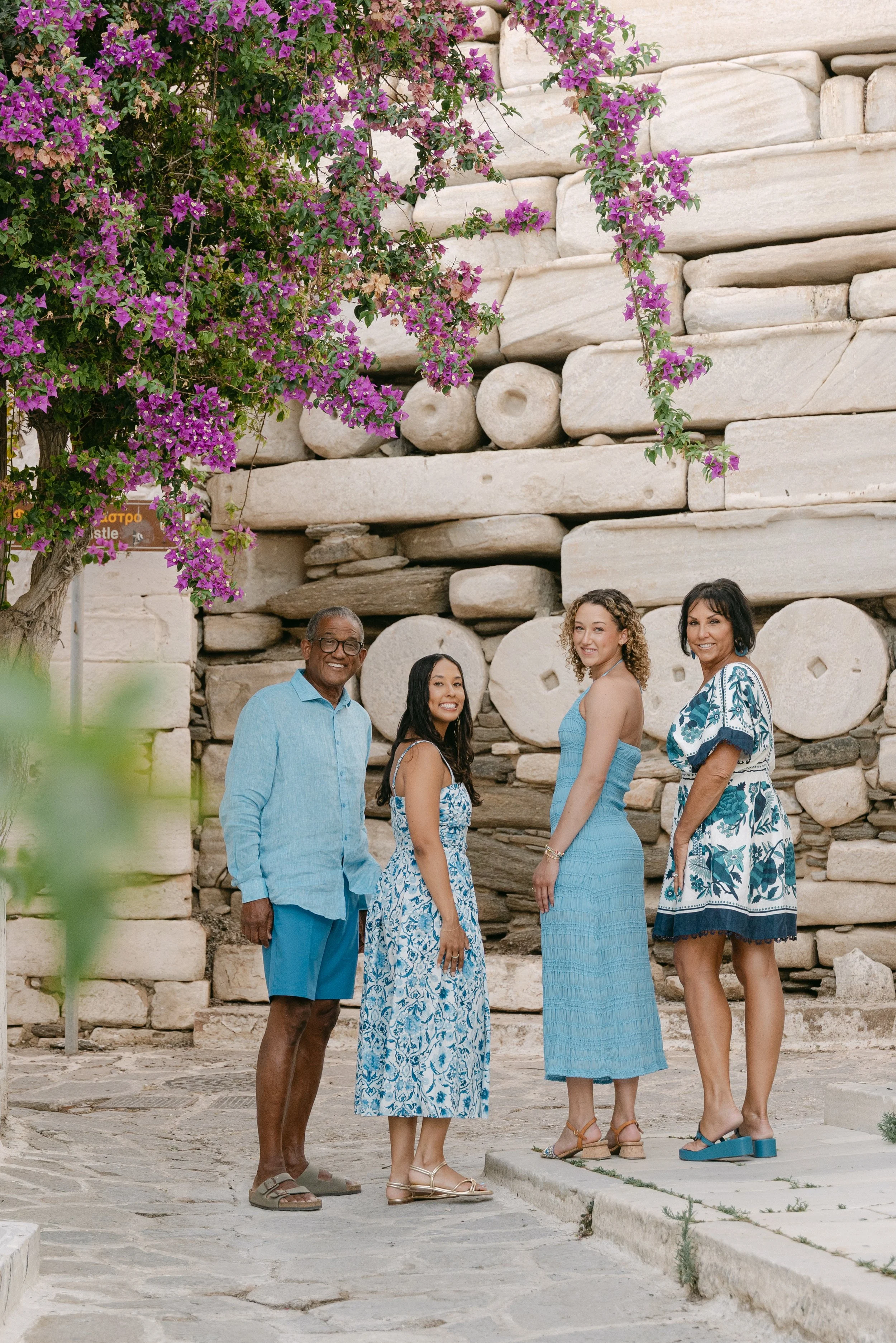 Four people standing in front of a stone wall with pink bougainvillea flowers overhead, all dressed in blue clothing.