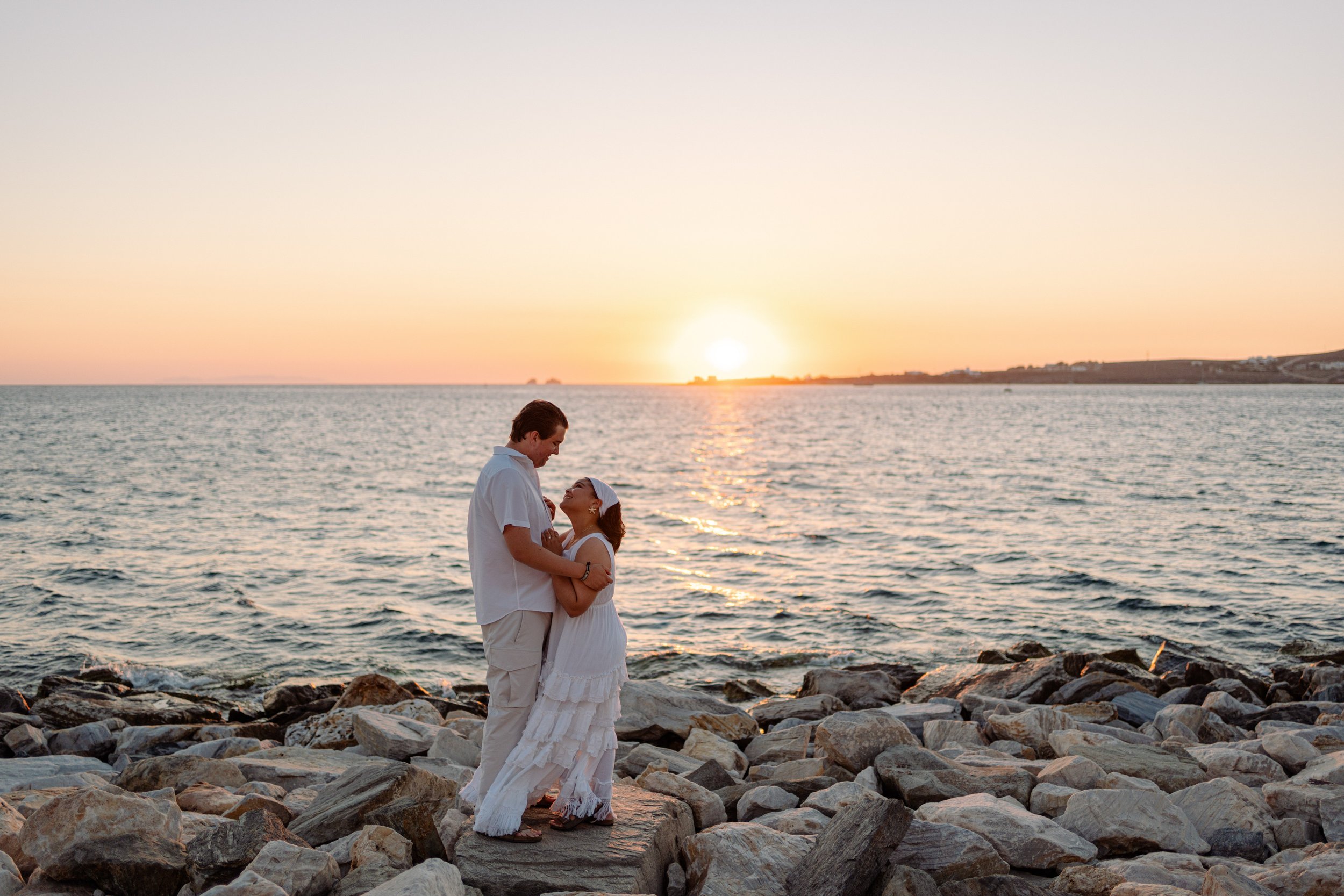 A couple stands on rocky shoreline during sunset, gazing into each other's eyes, with the ocean and sunset in the background.