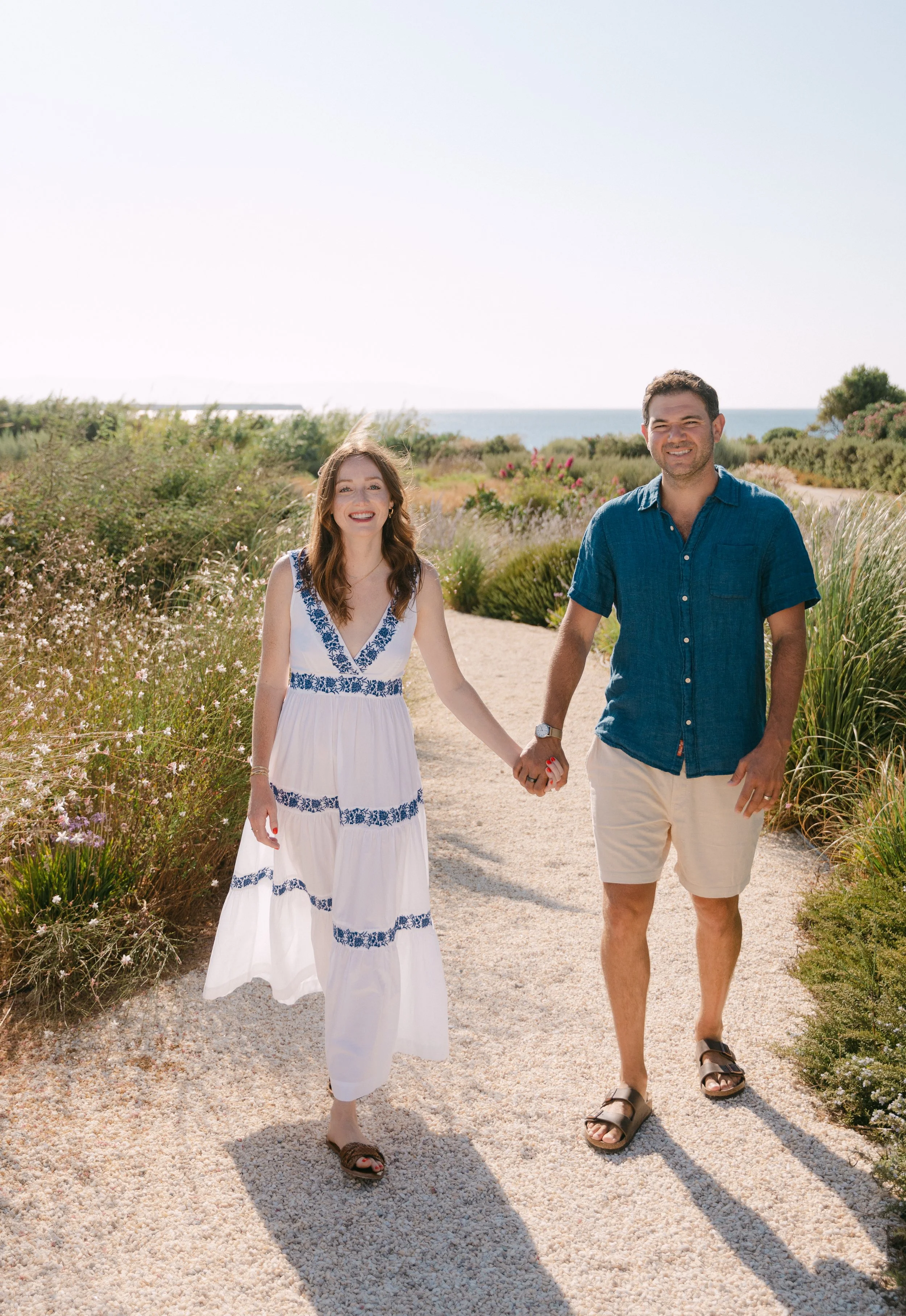 A smiling couple holding hands and walking on a sandy path surrounded by flowering shrubs, with the ocean visible in the background on a sunny day.