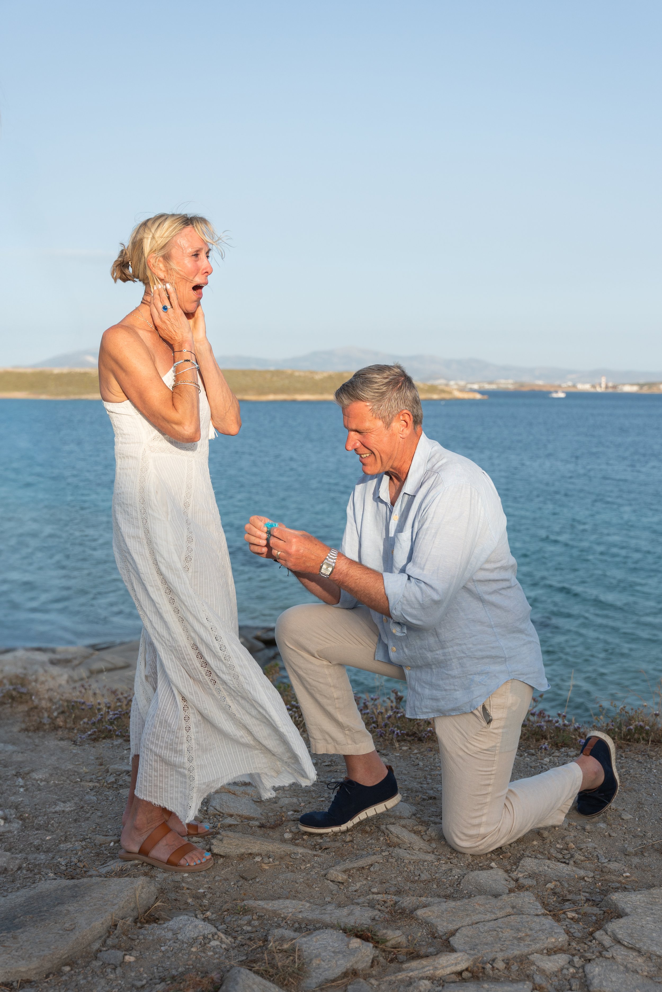 A man is proposing marriage to a woman by a body of water, with an emotional reaction from the woman.