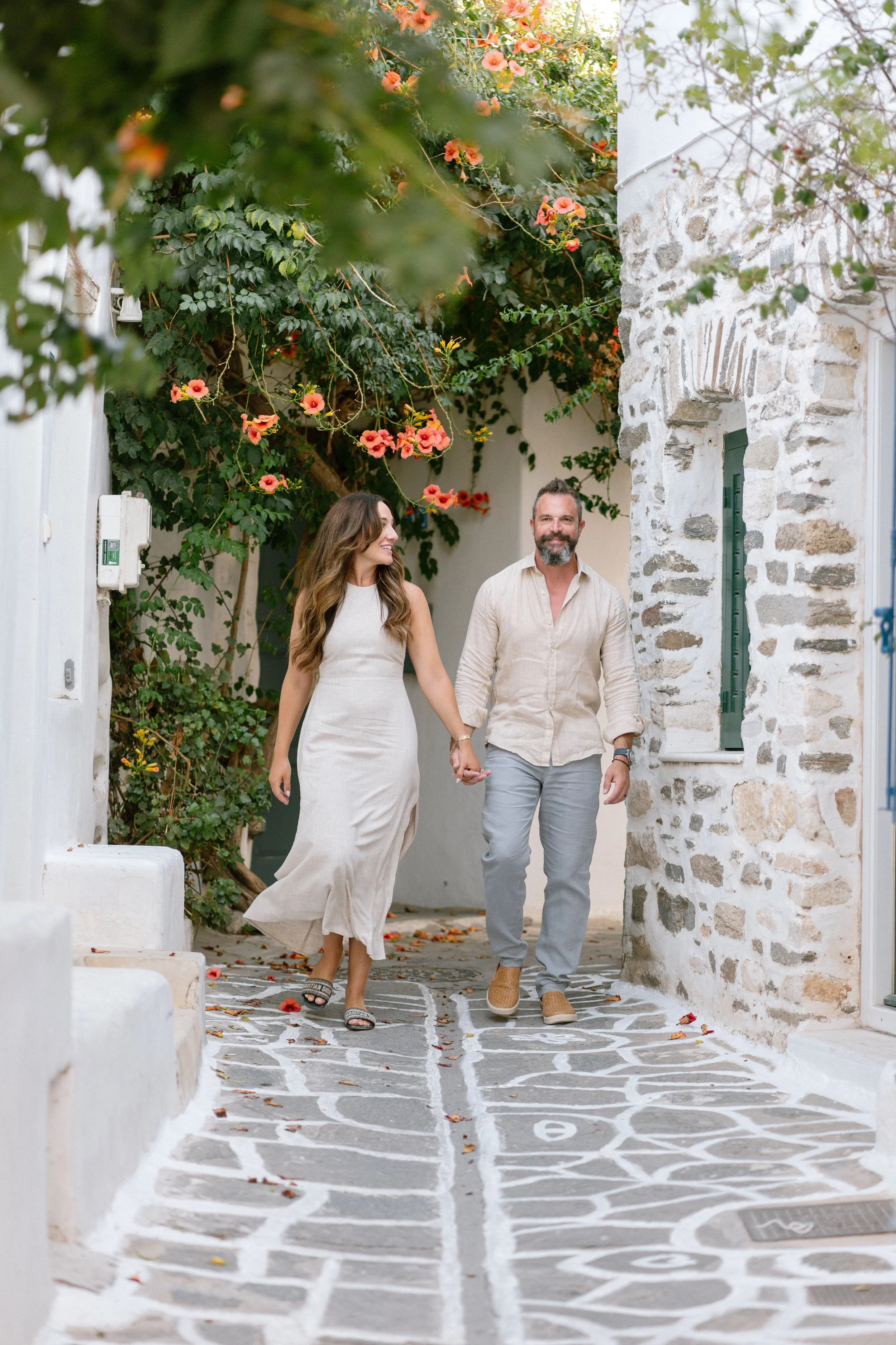 A couple holding hands and walking along a stone pathway with white painted lines and numbers, surrounded by white walls and blooming orange-pink flowers in a Mediterranean setting.