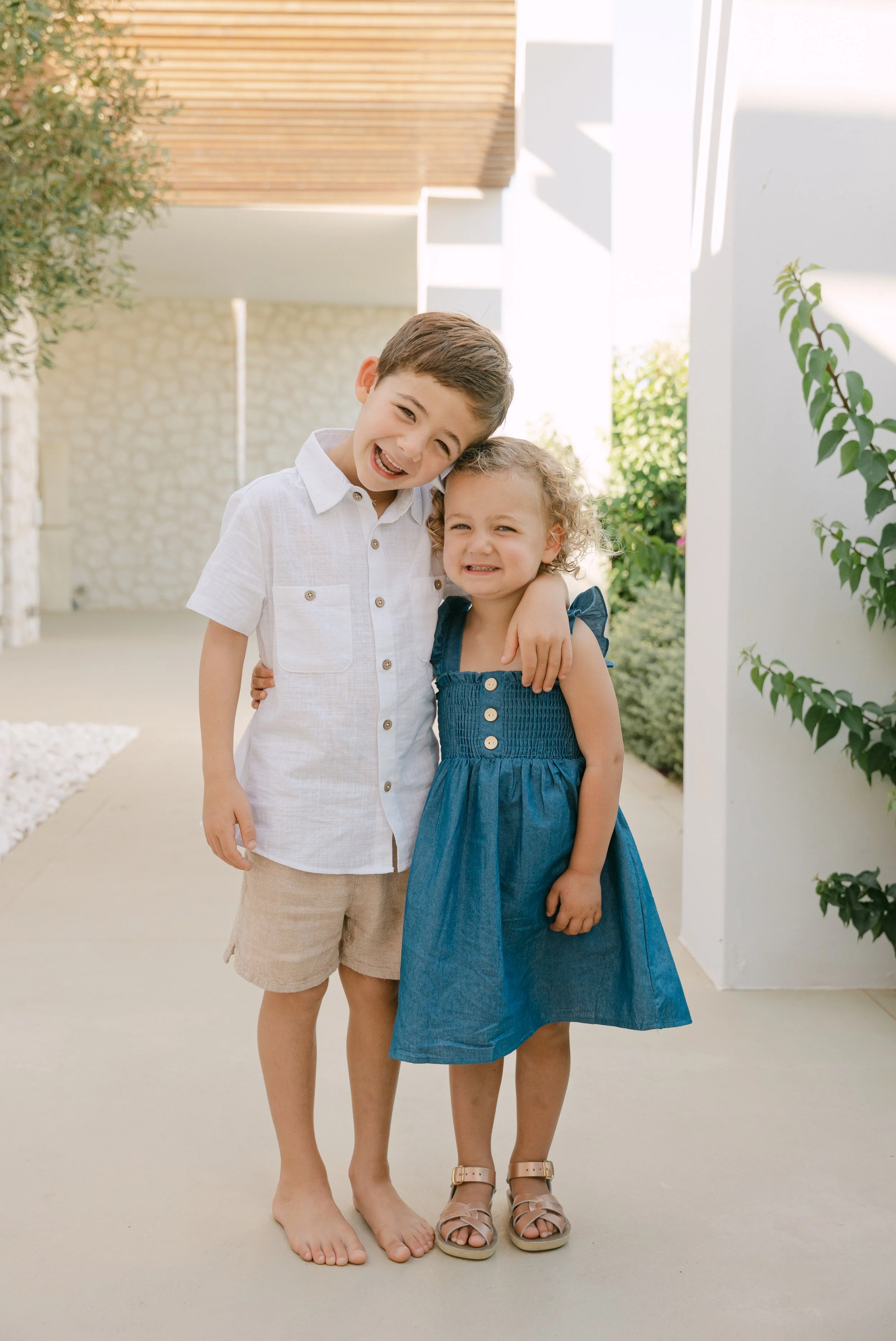 Two children, a boy and a girl, smiling and hugging outdoors near a white wall with greenery. The boy is barefoot, wearing a white shirt and beige shorts. The girl is wearing a blue dress and sandals.