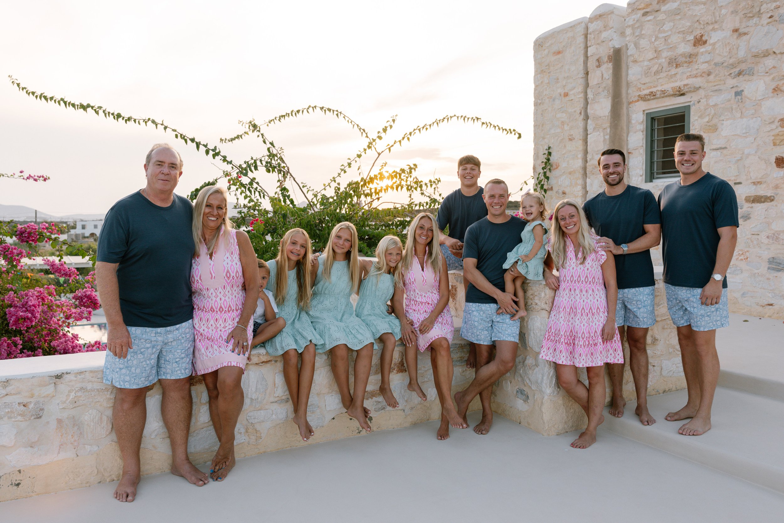 Family of fifteen posing outdoors on a stone ledge at sunset, wearing matching beachwear. The family includes children and adults smiling, with pink flowering bushes and a stone building in the background.